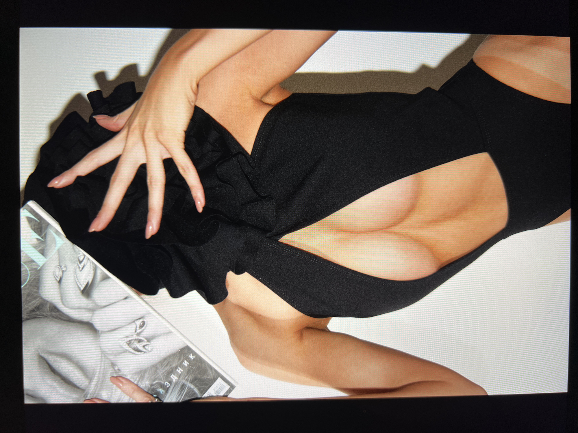 A woman is shown from the back, wearing a black swimsuit with a large cutout that exposes her breasts and upper back. Her head is covered by a black hooded garment with ruffles, and her hands are raised to cover her face. She is holding a magazine, part of which is visible. The background is a white wall. The image appears to be a photograph taken in a studio setting, with artificial lighting. There is no indication of a specific location in the city of Unknown, Russia.
