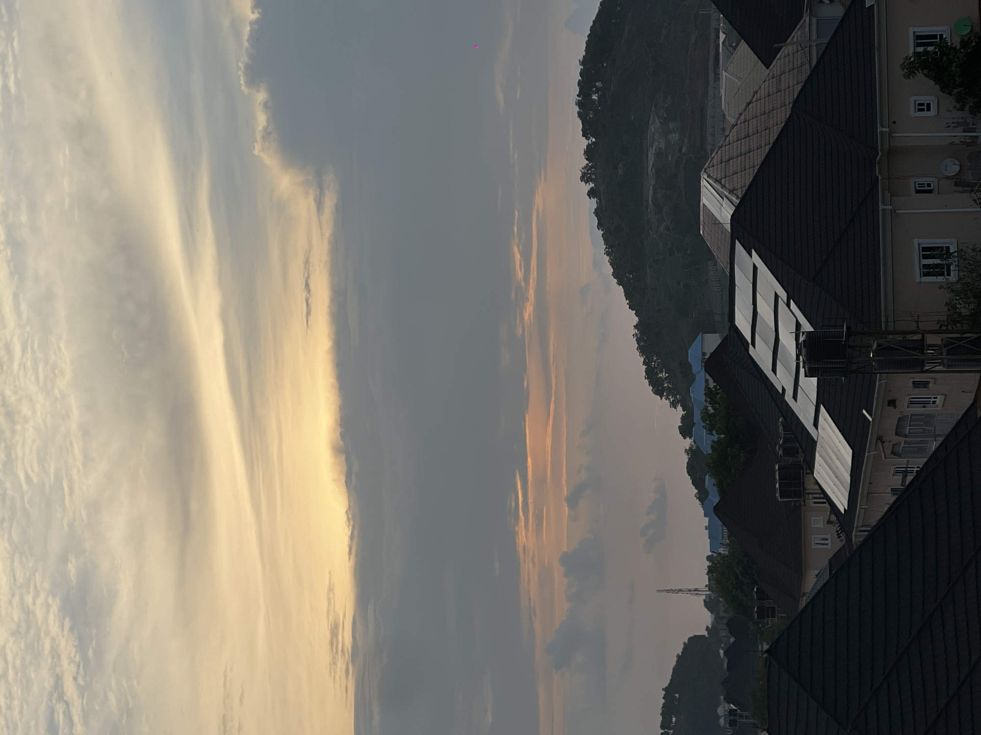 The image shows a cityscape at dusk in Abuja, Nigeria. In the foreground, several houses with dark, pitched roofs are visible. Some of these roofs have solar panels installed. In the mid-ground, more houses and buildings, some with light blue roofs, are nestled amongst trees. A large, green, tree-covered hill dominates the background. The sky is filled with a mix of gray and yellow clouds, suggesting either an approaching storm or the remnants of a sunset. A tall communication tower is visible on the left side of the image, further indicating an urban environment. The overall impression is one of residential development at the edge of a more natural, hilly landscape.