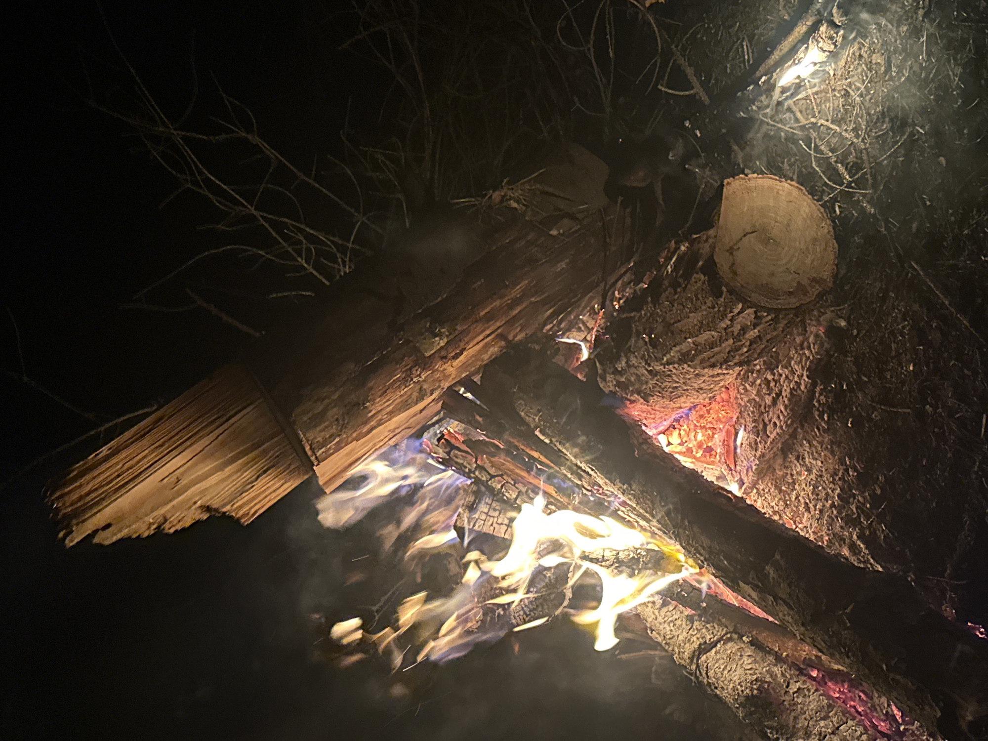 A night shot depicts a campfire with burning logs. The flames are yellow and white, with some red embers visible within the fire. There are dark tree branches visible in the background. The location is likely rural, though there is no confirmation that the image was taken in Юзуфоўскі сельскі Савет, Belarus.