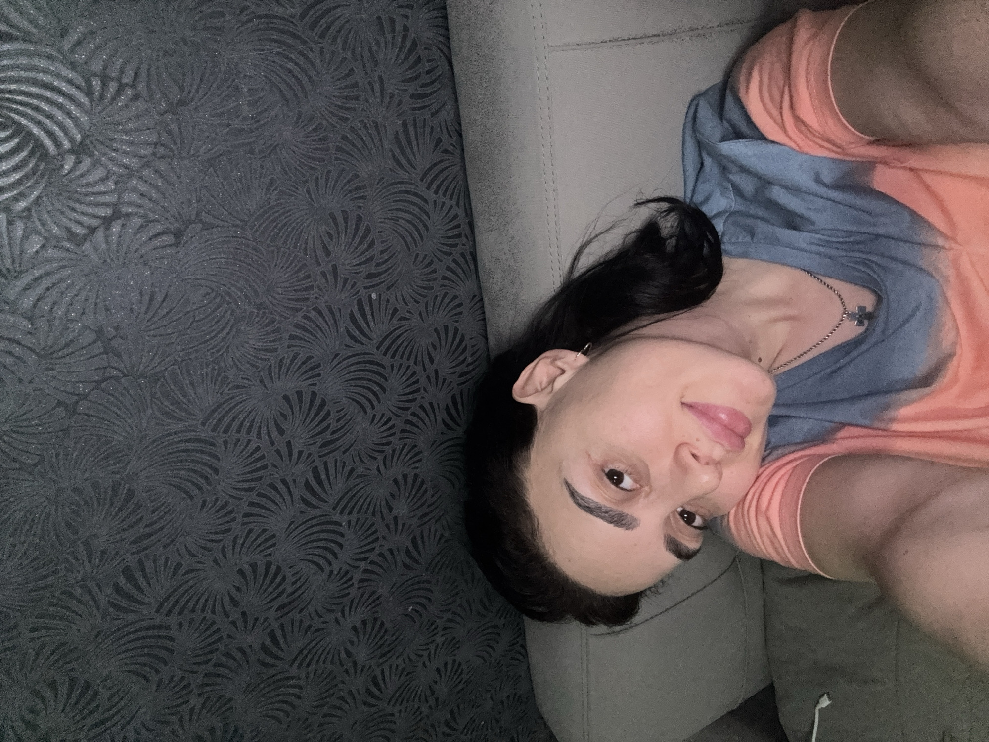A selfie photograph shows a woman lying on a grey couch with a dark grey patterned backdrop. She has dark hair, brown eyes, and is wearing a t-shirt that is peach and blue. She has a silver necklace with a cross pendant. A white headphone wire is visible near the bottom of the couch. There is no information to determine if this image was taken in Tbilisi, Georgia.