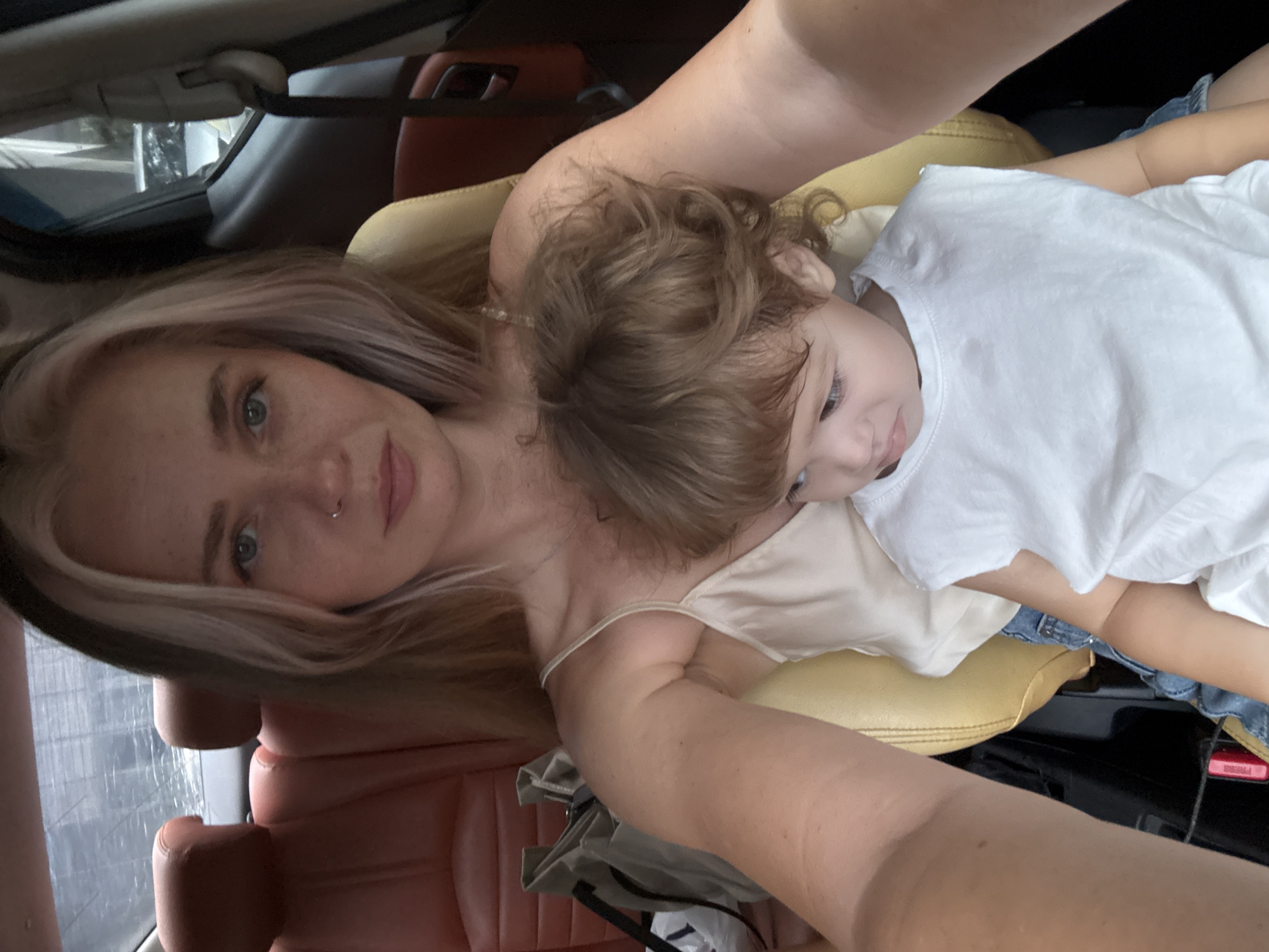 A woman with blonde and pink-streaked hair and a nose ring is taking a selfie in the driver's seat of a car, likely in Choeng Thale, Thailand. She is wearing a light-colored camisole. A young child, wearing a white t-shirt, is seated on her lap, facing away from the camera. The car's interior features reddish-brown leather seats. A light-colored tote bag is visible behind the woman. The background outside the car windows appears blurred and somewhat overcast.