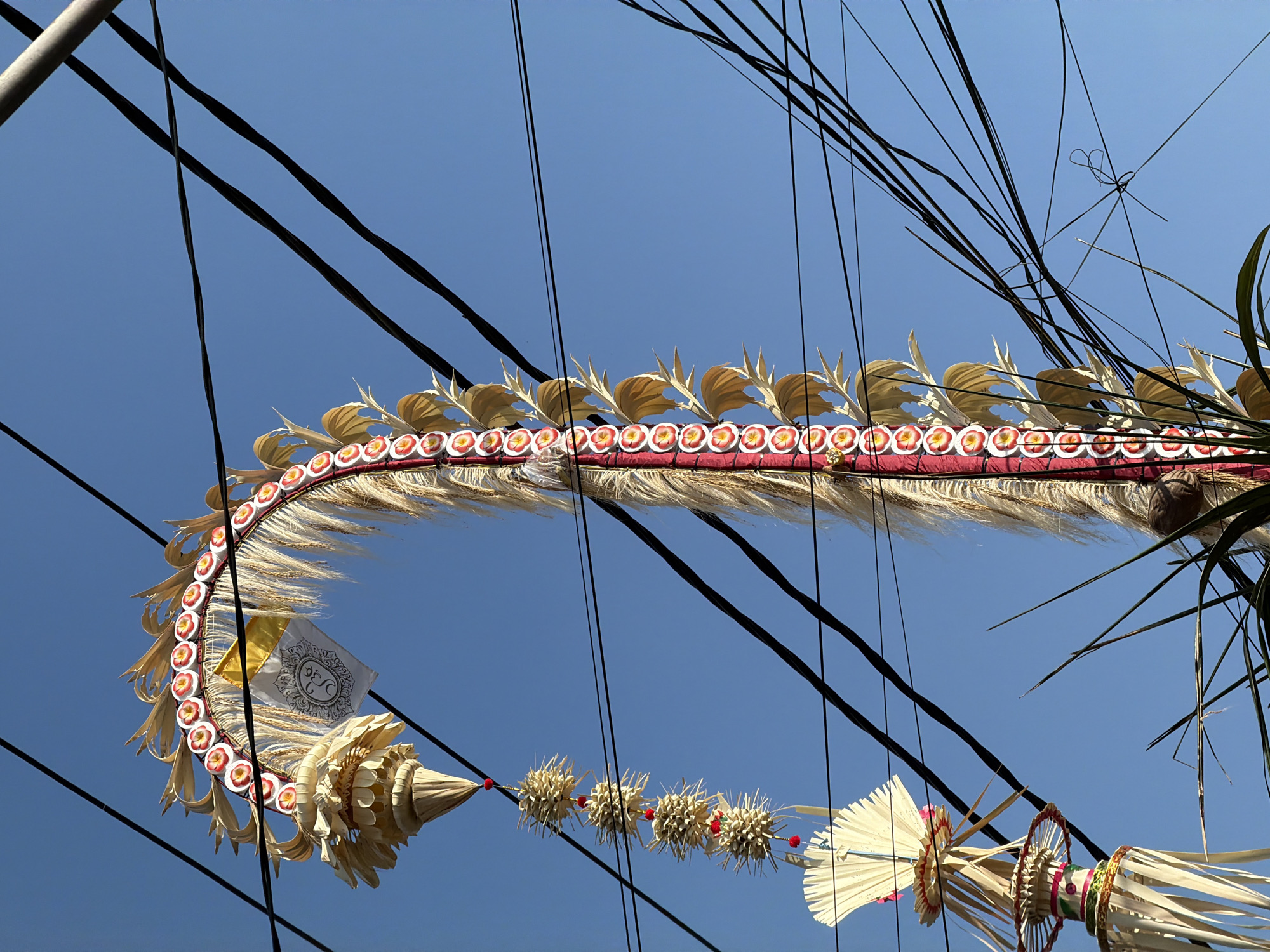 The image, taken in North Kuta, Indonesia, captures an ornate Balinese Penjor, a tall, bamboo pole decorated with various offerings and symbols, dominating the frame against a clear blue sky. The Penjor is adorned with a curved structure featuring a row of red and white floral-like ornaments and golden leaf-shaped accents. Hanging from it are several smaller, star-shaped woven decorations, each tipped with a red bead. The Penjor is partially obscured by a dense network of black electrical wires. In the lower left corner, another decorative element, possibly part of a different structure, is visible, crafted from woven palm leaves. Some green foliage protrudes into the lower part of the frame, adding a touch of natural greenery to the scene. The overall impression is one of cultural celebration and the juxtaposition of traditional artistry with modern infrastructure.