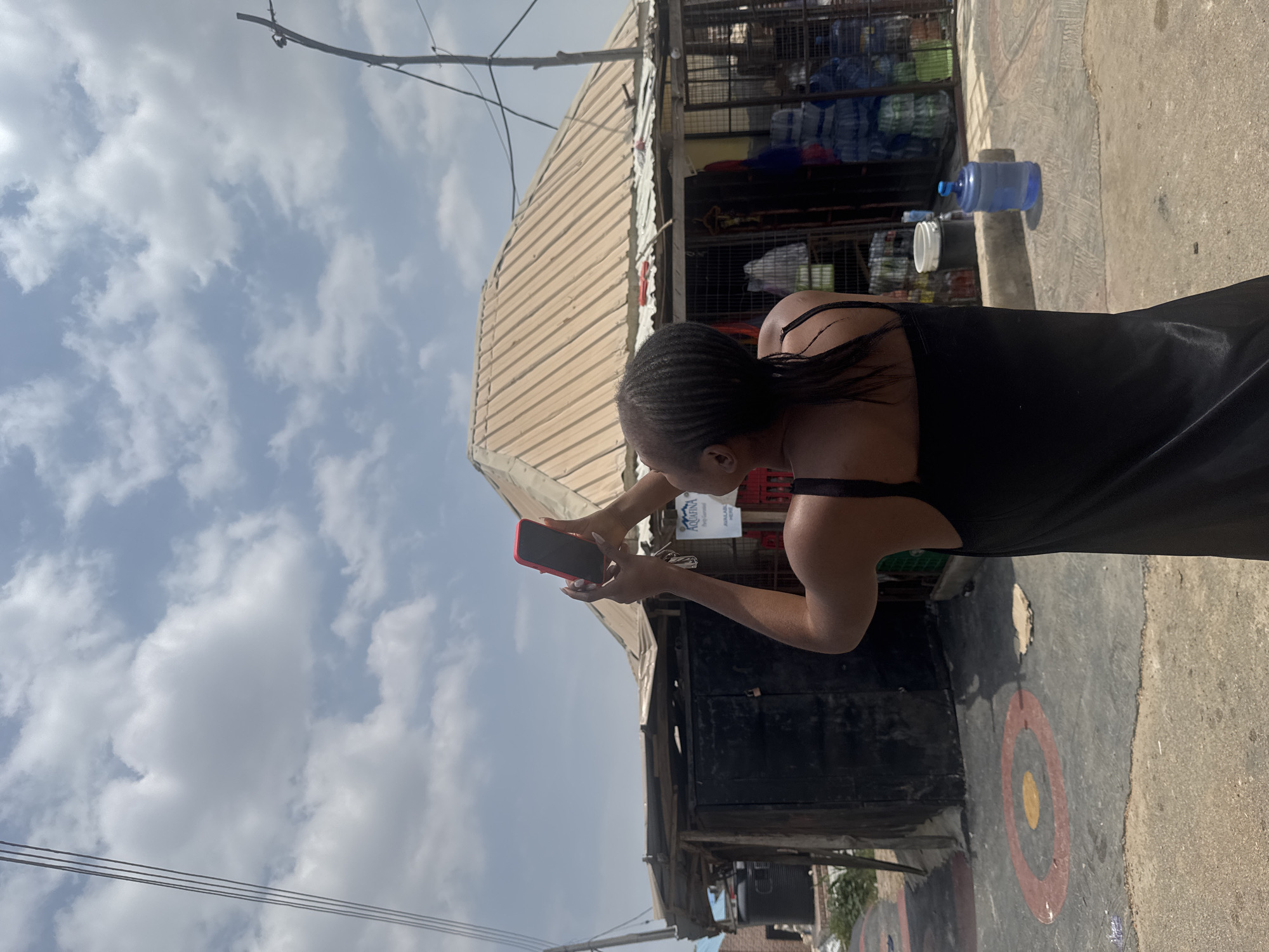 A woman with dark hair styled in braids is holding a red smartphone up to photograph the sky. She is wearing a black sleeveless dress. Behind her, a small shop with a corrugated metal roof is visible. The shop is stocked with bottled water and other goods, some of which are displayed on shelves behind a wire mesh. A large blue water cooler bottle stands in front of the shop. The ground in front of the shop is paved with concrete, and there are circular painted markings on the pavement. Utility poles and wires are visible against the cloudy sky. The scene appears to be an outdoor market or street vendor area in Abuja, Nigeria.