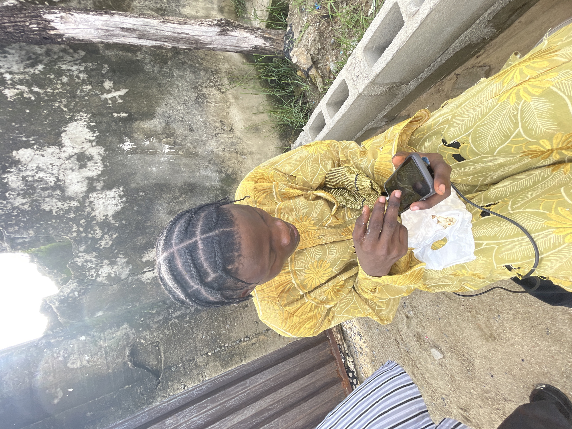 A medium shot captures a person seated outdoors, engaged with a smartphone. The individual, adorned in a yellow patterned garment, appears to be examining the device, with a white tissue nearby. The surroundings indicate an outdoor urban setting, with a wall and concrete blocks in the background. Vegetation is visible, suggesting the presence of greenery. The location is Ado, Nigeria.