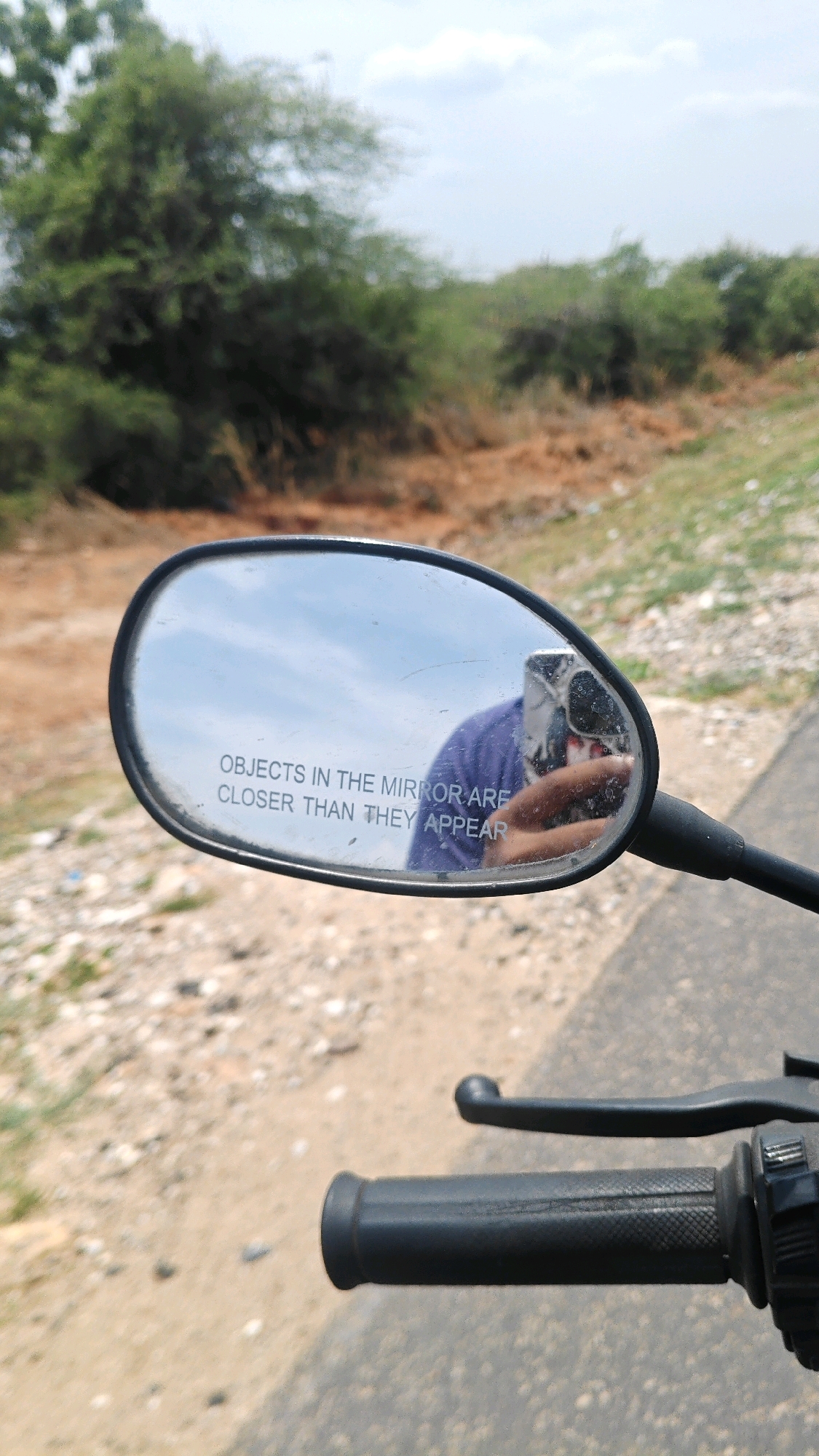 An outdoor, eye-level, close-up, vertical, medium shot captures a motorcycle's rearview mirror reflecting a person's purple shirt and hand holding a smartphone, as well as the surrounding environment. The mirror is oval-shaped with a black plastic casing and is mounted on a black arm. Text on the mirror reads "OBJECTS IN THE MIRROR ARE CLOSER THAN THEY APPEAR". The background is a blur of dry, brown earth and sparse green vegetation under a bright, hazy sky. The scene appears to be in Kotta Jadadevi, India, likely a rural or semi-rural area given the terrain and vegetation. The motorcycle's black handlebar grip is visible in the foreground.