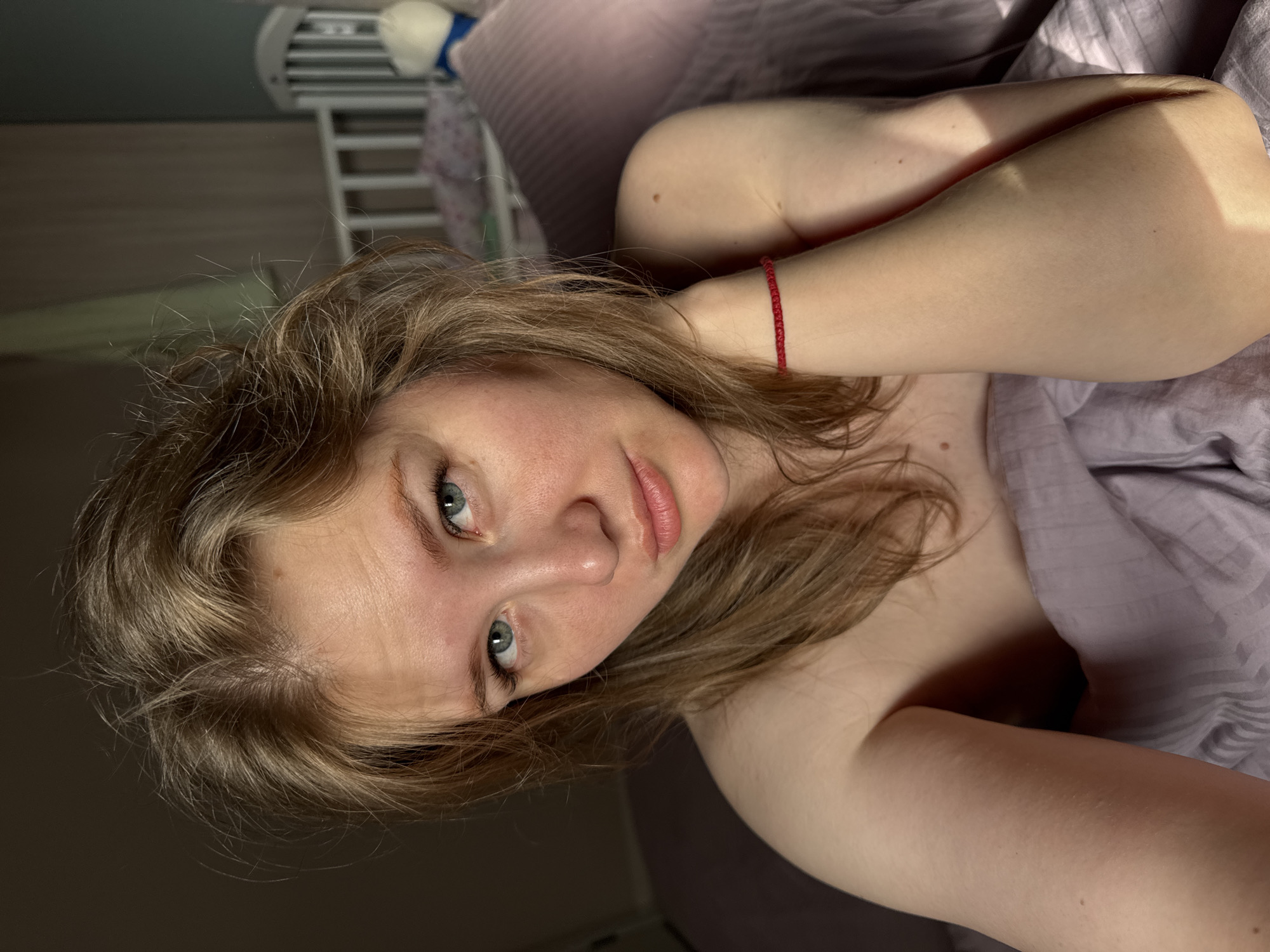This angled portrait shows a young person with light brown hair and blue eyes, lying down indoors. The individual looks directly at the viewer with a neutral expression. A thin red string bracelet is worn on their left wrist. Strong natural light illuminates the right side of their face and arm, creating distinct shadows and highlights, indicative of morning or late afternoon sun. The rest of their body is partially covered by a light purple, striped blanket or sheet. In the blurred background, elements of a bedroom are visible, including a white slatted structure (possibly a headboard or bed frame) and a pink cushion. The scene suggests a candid moment, likely in a bed. The photo was taken in Ферма, Russia.