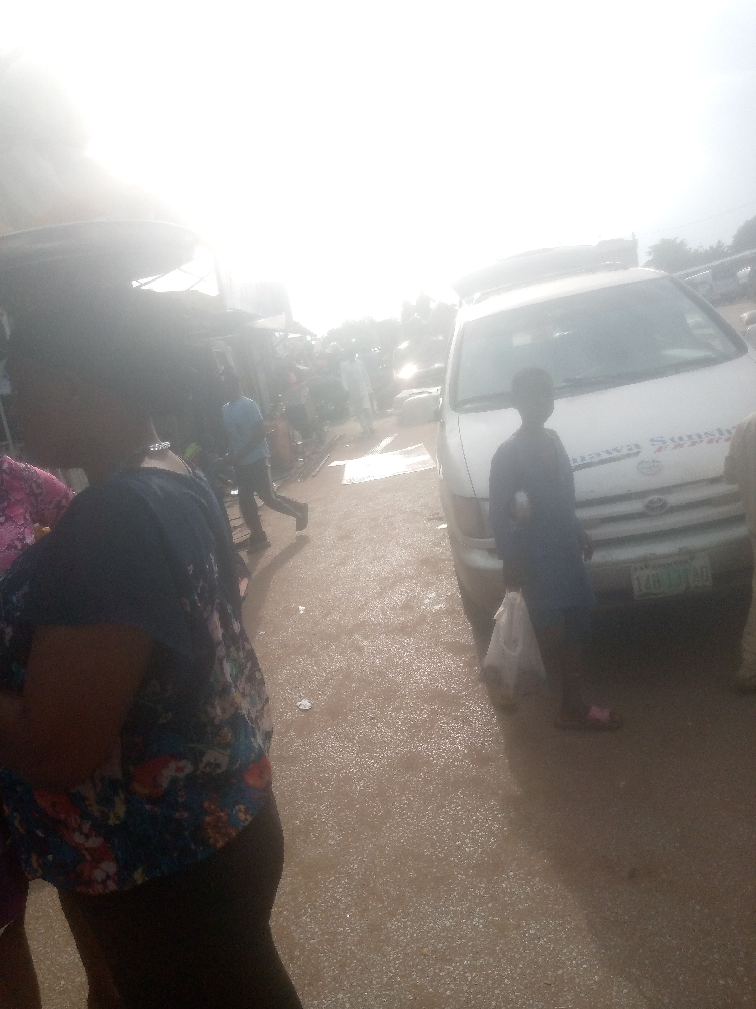 In Jimeta, Nigeria, a busy street scene unfolds under a bright, hazy sky, possibly indicating early morning or a dusty atmosphere. In the foreground, a woman, partially obscured by the camera's angle and bright light, is visible from the shoulders down, wearing a dark blue top with a vibrant floral pattern. To her left, another person, also partially visible, wears a pink floral garment.
The central focus of the image is a white minivan parked on a wet, dirt road. The minivan has text on its hood, partially visible as "Guamawa Sunshine Express". A young boy, dressed in a dark blue tunic and trousers, stands beside the van, holding a white plastic bag. His feet are clad in pink sandals. The minivan's license plate is clearly visible as "148-13-TAO".
In the background, a blurred crowd of people are seen walking and standing. Some appear to be vendors or shoppers in a market-like setting, with stalls or structures visible behind them. The overall impression is one of a dynamic, public space in motion, typical of a market or transport hub. The wet ground suggests recent rain or a deliberately watered area, common in hot climates to control dust. The bright sky creates a strong backlight, washing out some details and giving the image a slightly diffused, almost ethereal quality.