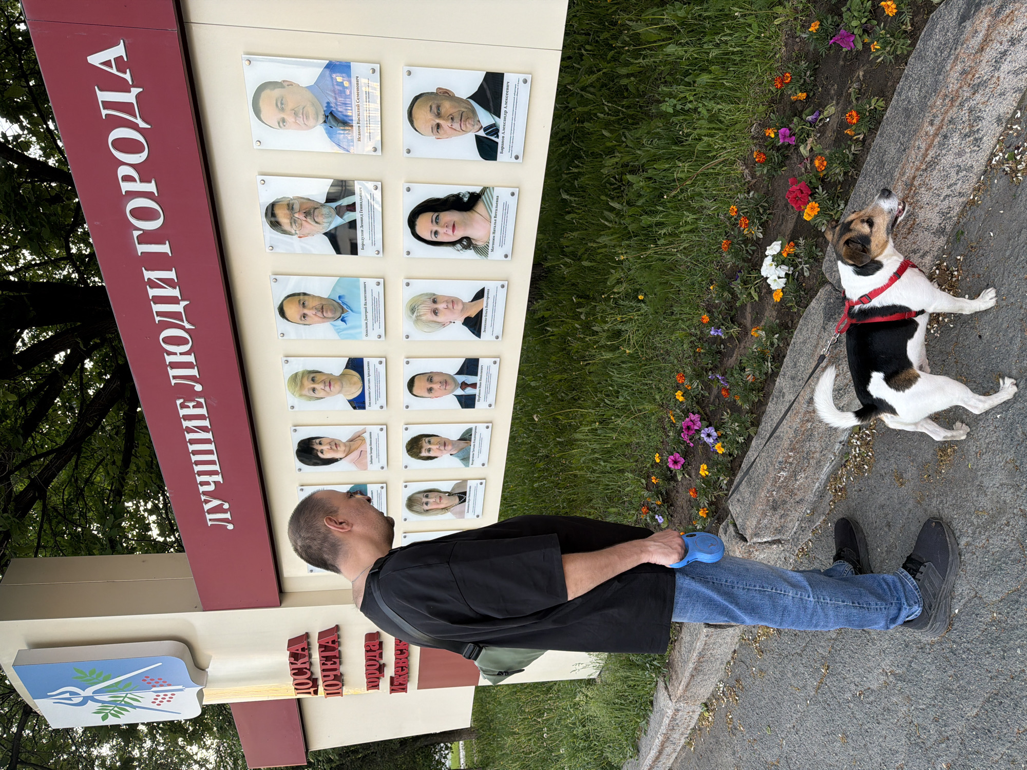 The image shows a man and a dog standing near a memorial board, likely located in Izhevsk, Russia, which features photographs of distinguished citizens. The board is labeled "Лучшие люди города" (The Best People of the City) and includes a coat of arms and the words "ДОСКА ПОЧЕТА города Ижевск" (Izhevsk City Board of Honor). The dog, wearing a red harness, stands on the edge of a stone path, near a flower bed with colorful flowers. The man, wearing a black shirt, blue jeans, and black shoes, appears to be looking at the memorial board. The photograph appears to be taken during the day in a park-like setting with trees and greenery visible in the background.