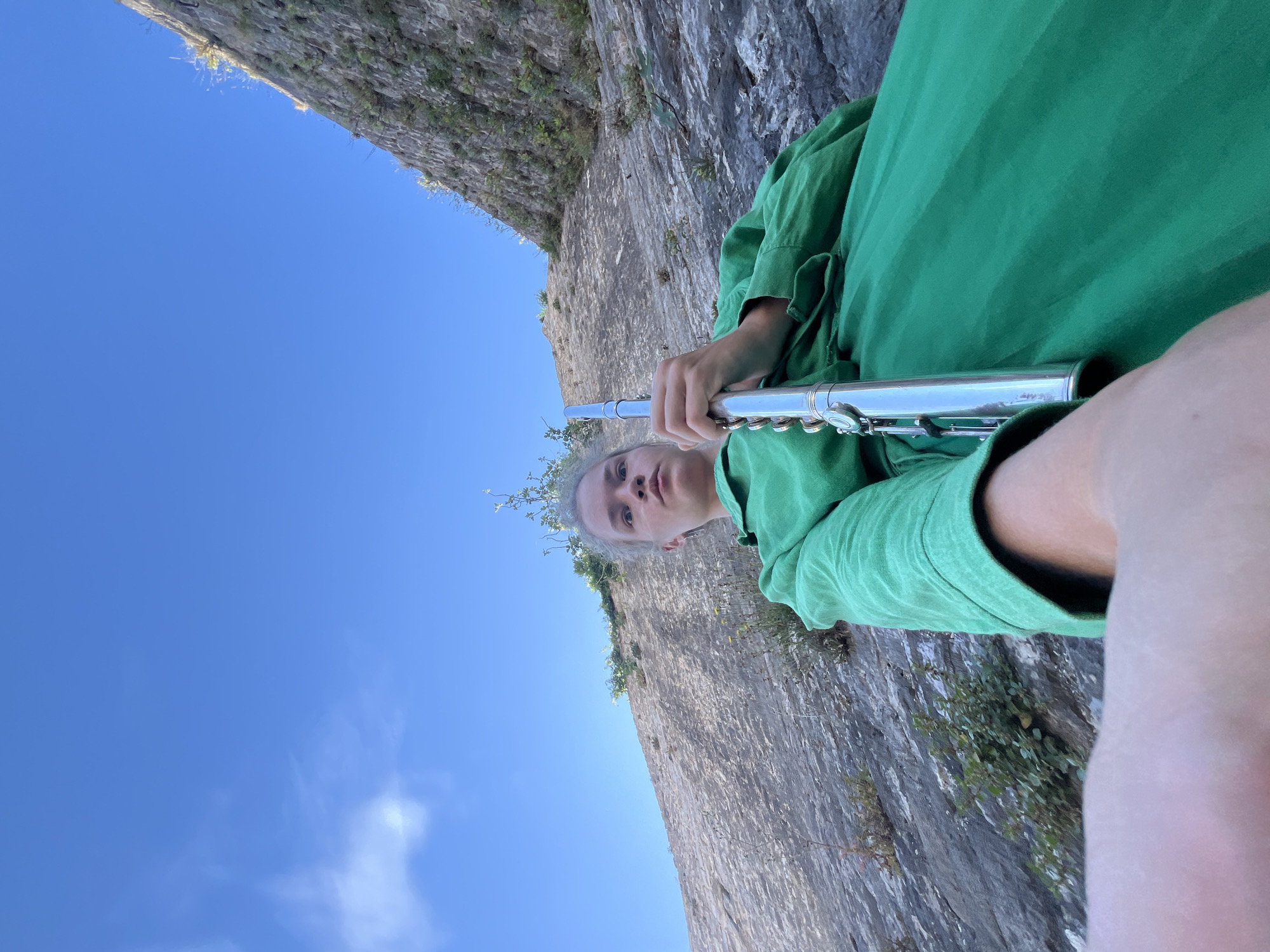 A low-angle, wide shot shows a person in a bright green dress holding a silver flute against a rugged, rocky hillside under a clear blue sky. The person's face is partially visible, looking towards the camera with a surprised expression. The flute is held horizontally across the person's lap, with the mouthpiece end closer to the left of the frame. The rock face behind the person is textured and appears to be a natural formation, possibly part of the mountainous terrain surrounding Budva, Montenegro. Sparse vegetation, including small green plants and yellow wildflowers, grows from crevices in the rock. The sunlight casts shadows and highlights on the rock face, indicating a bright day.