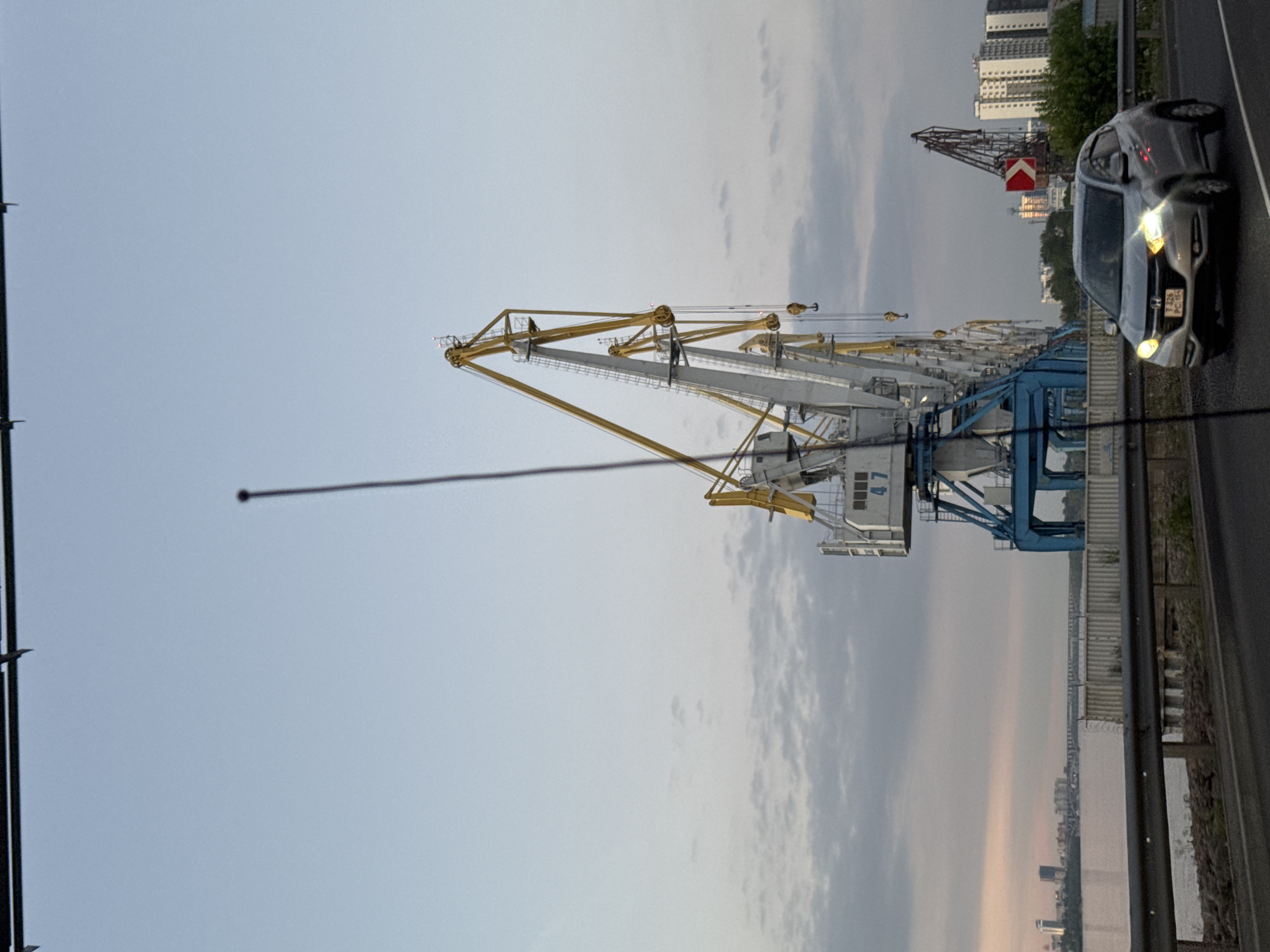 The image captures a waterfront scene in Krasnoyarsk, Russia, likely at the port. Dominating the frame are several large industrial cranes, one with the number "47" visible. These cranes, with their white and yellow structures, suggest active cargo handling operations. In the foreground, a silver car, possibly a Honda HR-V, is driving on a road. The car's headlights are illuminated, indicating it might be dusk or an overcast day. A body of water, likely the Yenisei River, stretches across the middle ground, with a city skyline and a bridge visible in the distance on the left. The sky is partly cloudy, with soft, diffused light. A thin, dark vertical line, possibly an antenna or a wire, runs through the center of the image, slightly obscuring the view. The overall atmosphere is industrial and functional, with hints of urban life and natural landscape.