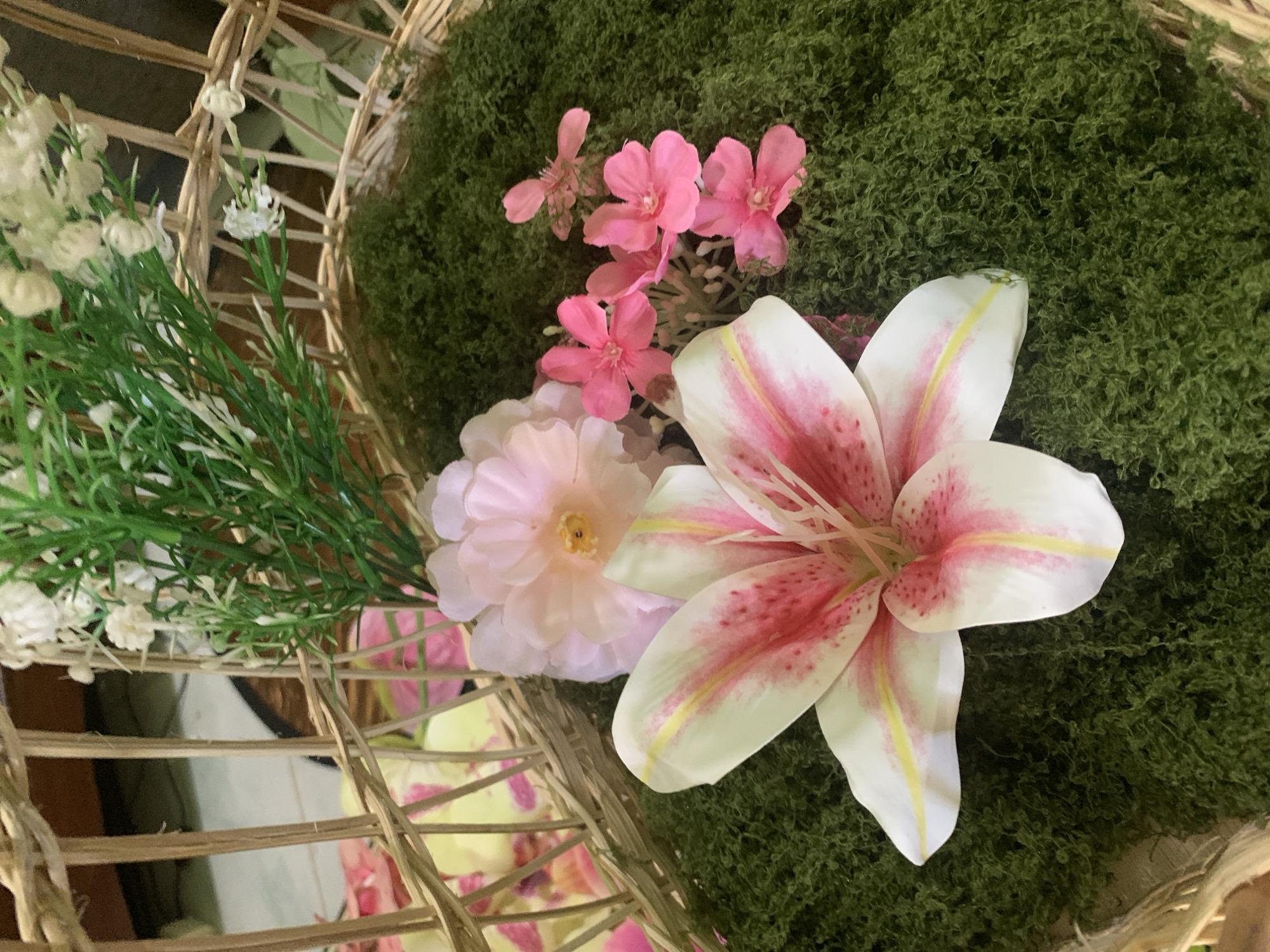 The image displays a close-up view of a decorative arrangement within a woven basket. The basket is filled with green, moss-like material. Prominently featured is a large, white lily with pink and yellow accents. To the right of the lily are several small, bright pink flowers. Behind the lily and to the left, a larger, pale pink flower is visible. At the top of the image, green artificial foliage with small white flowers emerges from the basket. The background is blurred, showing what appears to be part of the basket's weave and some colorful decorative elements. There are no people or animals present in the image. The location is not identifiable as Kuala Pembuang, Indonesia, based solely on the visual information provided.