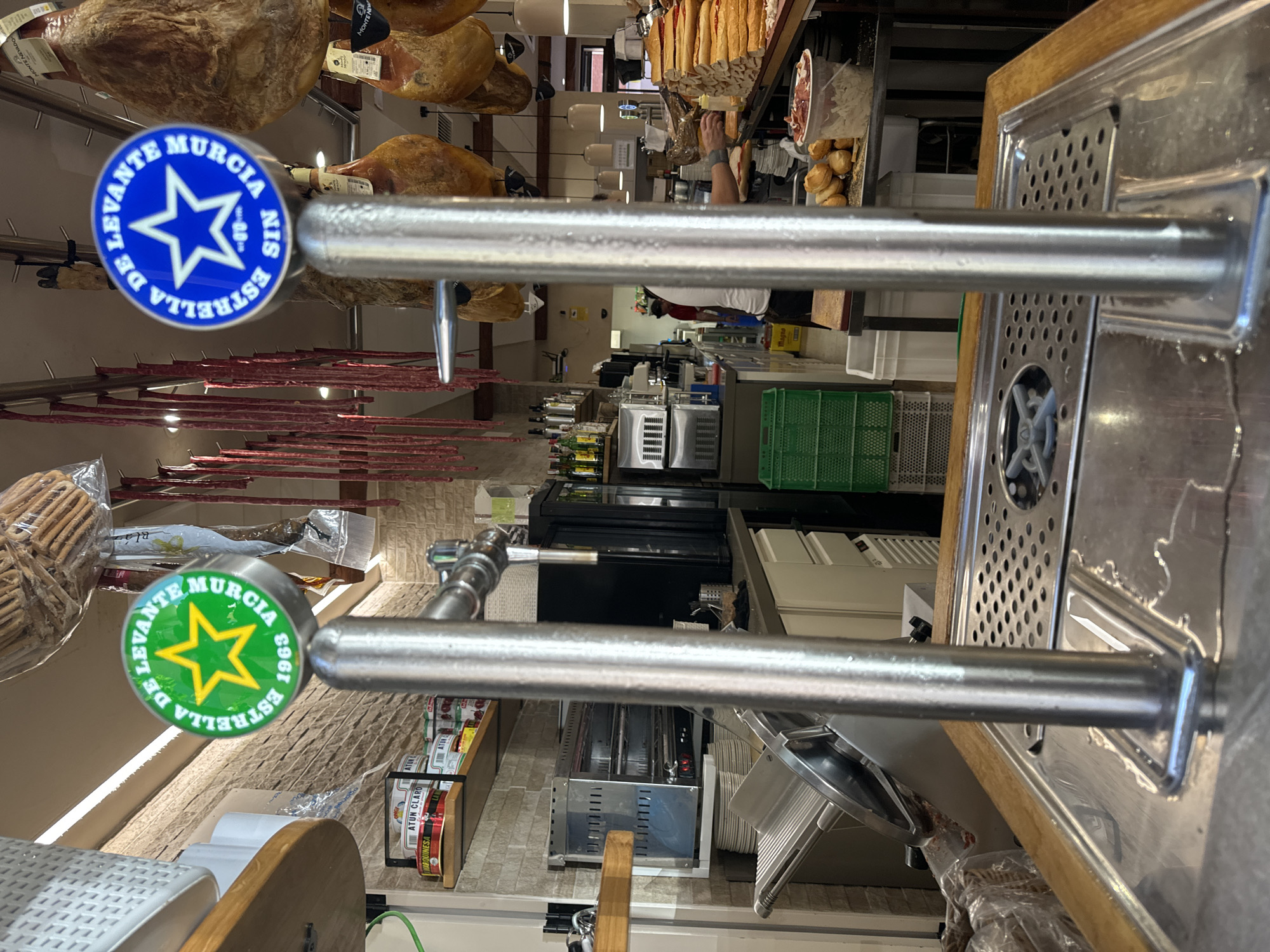 This media file, likely taken in Murcia, Spain, provides a close-up view of a bar or restaurant counter, focusing on beer taps and various food items. The scene appears to be indoors, possibly a market or a specialty food store, given the presence of cured meats, bread, and canned goods like tuna.
The prominent subjects are two beer taps, each adorned with a circular blue and green sticker. The stickers feature a star design and text that reads "ESTRELLA DE LEVANTE MURCIA" and "6961". One tap is more in focus, showcasing the shiny metal of the tap and the detailed design of the sticker.
In the background, a variety of food items are visible. Large cuts of cured meat, possibly jamón, hang from the ceiling. Shelves display rows of canned tuna, some with labels reading "ATUN CLARO MARQUINESA" and "ATUN MAGNO". There are also loaves of bread and other food items that suggest a culinary setting.
The lighting suggests it could be daytime, with artificial lights illuminating the counter area. The overall atmosphere seems to be that of a bustling food establishment. There are no visible people clearly interacting with the camera, but the presence of food preparation areas and stocked shelves implies ongoing business. The overall impression is one of a well-stocked food vendor, with an emphasis on local products like Estrella de Levante beer and regional cured meats.