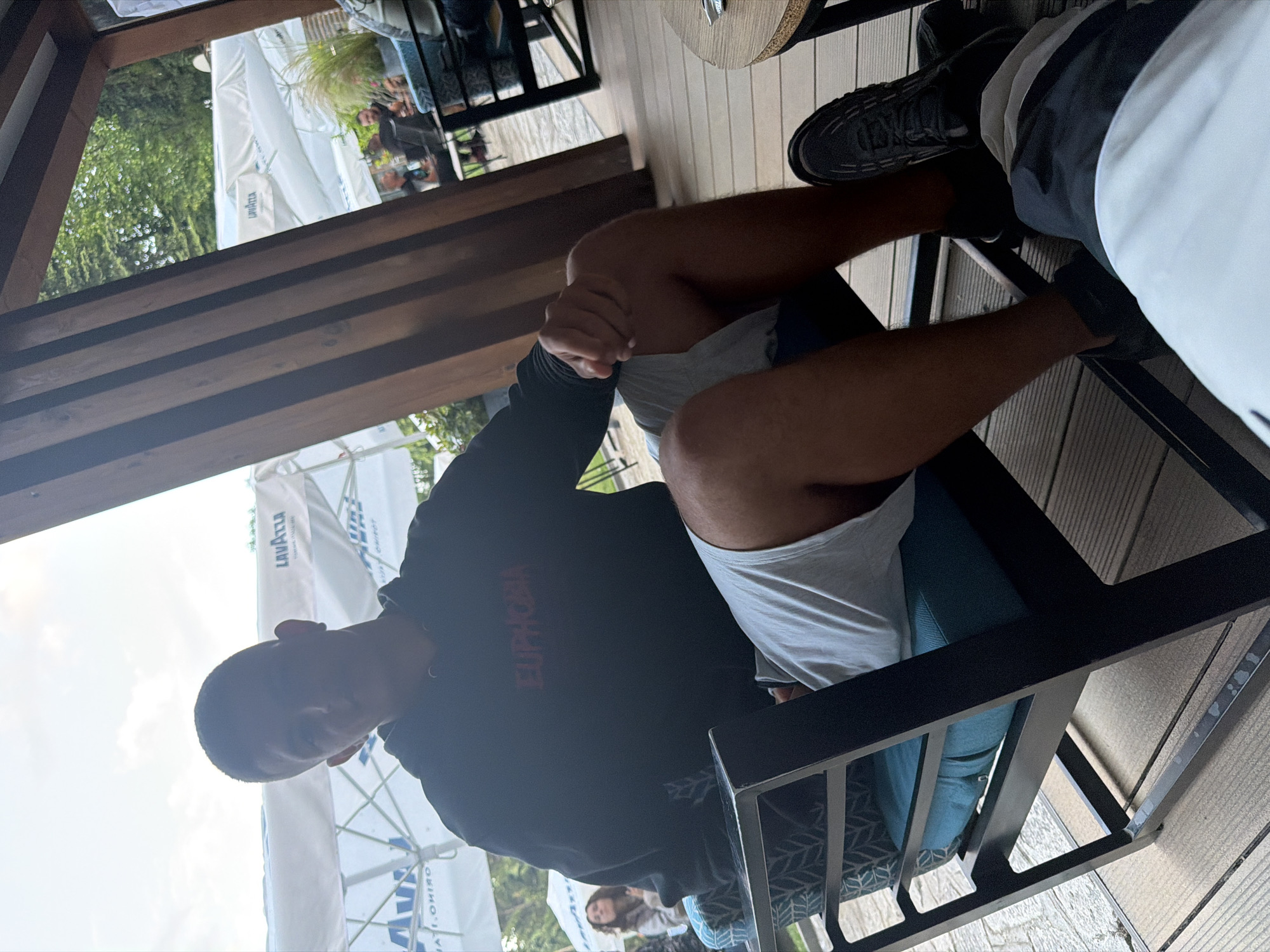 The image captures an outdoor or semi-outdoor cafe setting in Razlog, Bulgaria, under bright daylight. A person is prominently featured in the foreground, seated in a black metal-framed chair with a blue cushion, viewed from an oblique, slightly elevated angle. They are wearing a black hoodie with the word "EUPHORIA" printed in red on the chest, and light-colored shorts or boxers. Their hands are clasped, and their dark-skinned legs are visible. They are wearing dark-colored sneakers.
To the right of the main subject, another person's legs and feet are visible, wearing sneakers and athletic-style clothing in white and dark colors, suggesting they are also seated nearby. The flooring is a light-colored wooden deck. Overhead, dark wooden beams span across, hinting at a pergola or a covered seating area.
In the background, out of focus, large white outdoor umbrellas are visible, with "LAVAZZA" and "TORINO" (partially seen as "ORINO") printed on them, indicating a coffee brand. Green trees and other blurred figures of people seated at tables are also discernible in the distance. The scene suggests a relaxed social gathering or a break at an outdoor eatery.