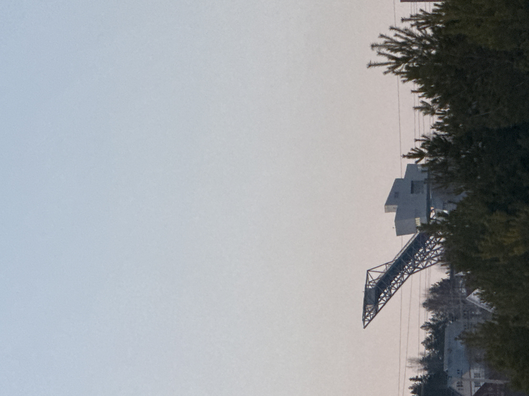 The image shows a pedestrian bridge in the city of Прикамский, Russia. The bridge has a metal framework and connects to a building. Trees are visible in the foreground. The sky is overcast.