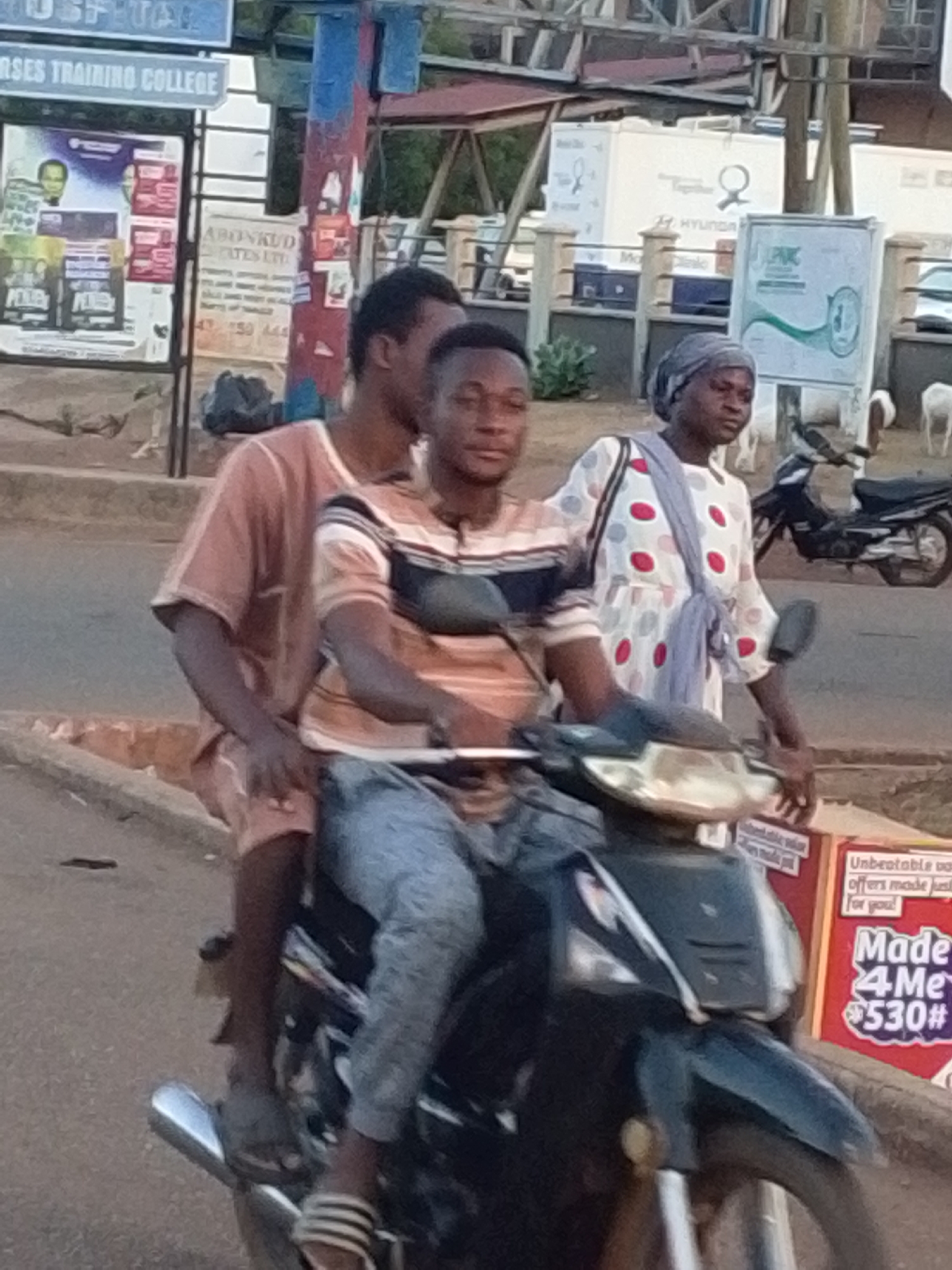 Here's a detailed description of the image:
**Scene Overview:** The image appears to be taken in Tamale, Ghana, likely on a city street. The backdrop features buildings and signage characteristic of an urban environment.
**Subjects & Actions:** The primary focus is on three individuals riding a black motorcycle/scooter.
* The driver is a young man wearing a striped shirt and jeans. He is looking directly at the camera.
* Behind him is another young man wearing a reddish-brown outfit. He is seated on the motorcycle as a passenger.
* To the right, standing beside the motorcycle on the side of the road, is a woman wearing a headscarf and a patterned white dress with red and blue polka dots. She is carrying a scarf.
**Objects:**
* A black motorcycle/scooter is the main mode of transportation.
* There are several signs visible in the background, including those for "NURSES TRAINING COLLEGE", "ABONKUD CATES LTD", and "Hyundai".
* A red cardboard advertisement with the words "Made 4 Me" written on it is to the right of the scooter.
**Environment:**
* The image is taken in broad daylight.
* The street is paved and shows signs of typical urban wear.
* Other motorcycles are parked nearby.
* Buildings and various advertisements line the street.