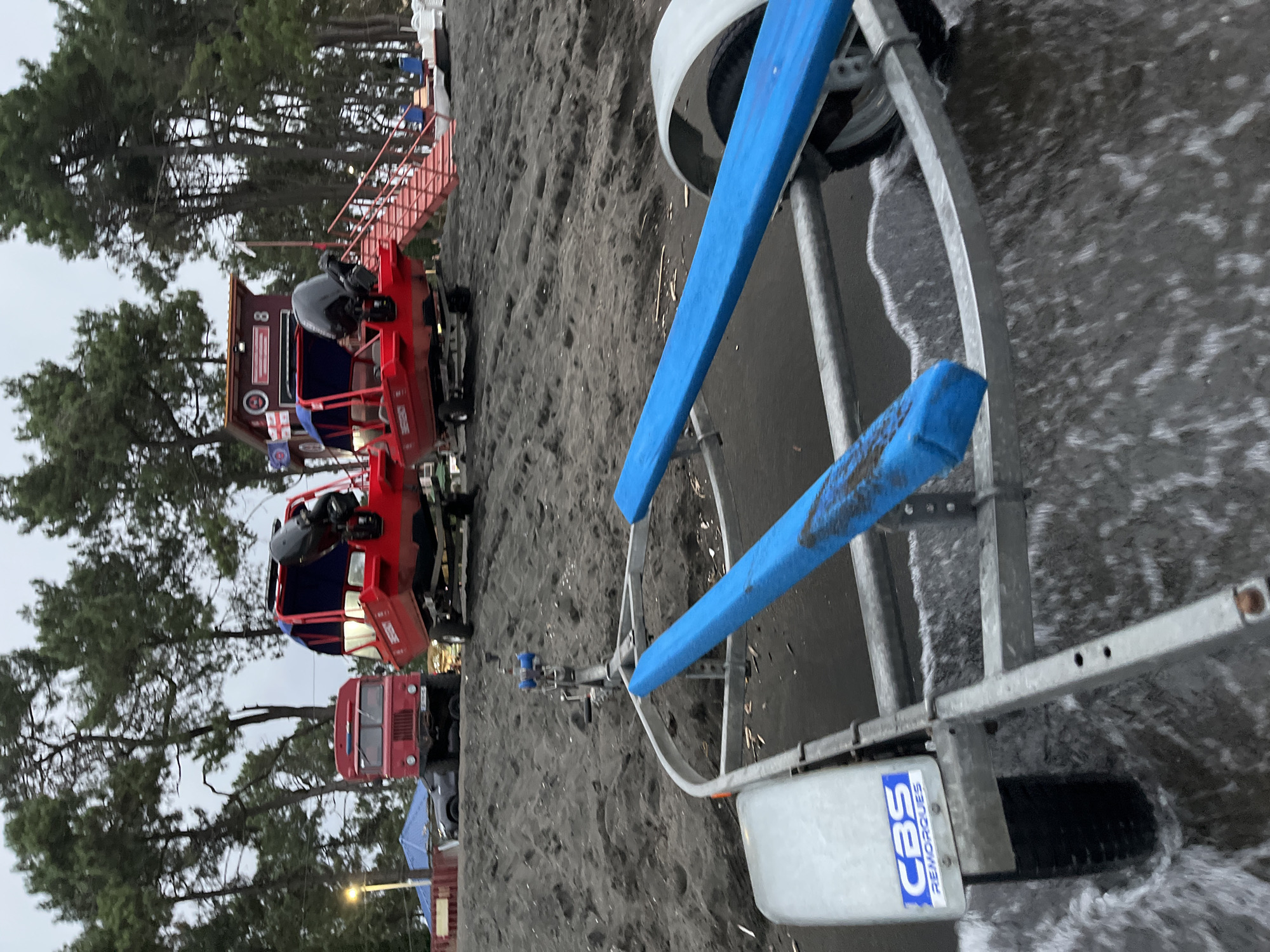 This image depicts a scene on a dark sand beach in Ureki, Georgia, likely near the Black Sea. In the foreground, a boat trailer with blue bunks and a "CBS REMORQUES" decal is partially submerged in shallow water, with ripples indicating movement. In the background, two red emergency response boats are visible on trailers. One boat, closer to the camera, has "EMERGENCY" written on its side. Behind the boats, a red truck is parked, and further back, a lifeguard station with red stairs and flags is present. Tall pine trees line the edge of the beach. The sky is overcast, suggesting a cloudy day. No people are visible in the image.