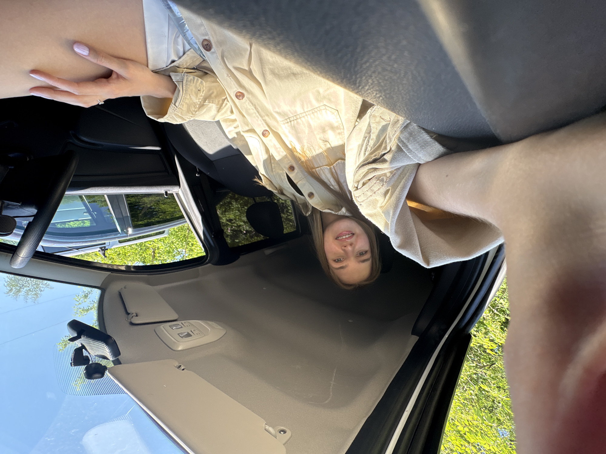 A woman with light-colored hair is photographed inside a car from a high angle. She is wearing a light-colored button-down shirt. Her left hand is visible near the top left corner of the frame, suggesting she may be holding the camera. The car interior is visible, including parts of the dashboard, steering wheel, and seats. Outside the car, through the windows, green trees and foliage can be seen, indicating a natural or park-like environment. There is no information to determine the location in the city of Perm, Russia.