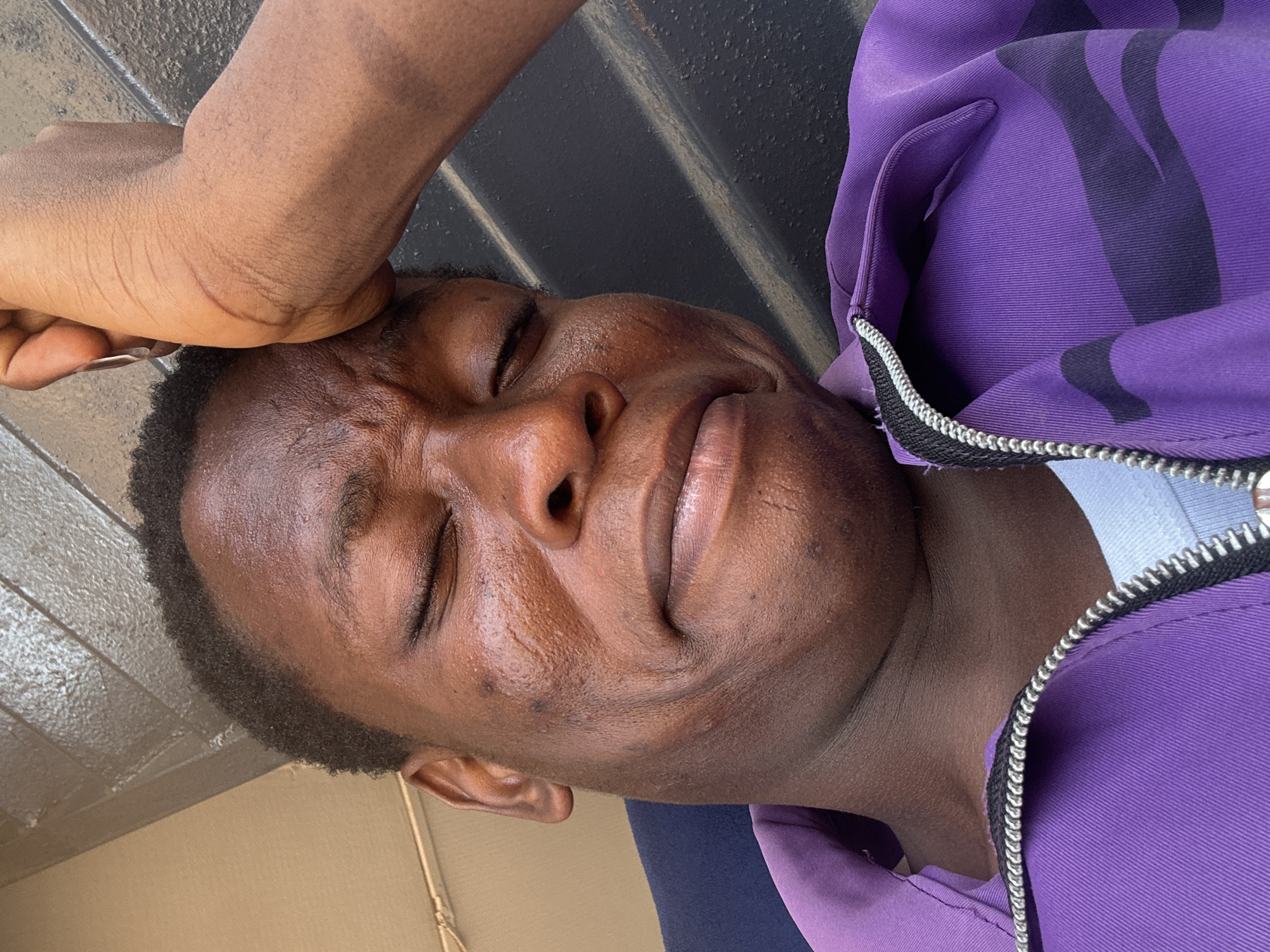 A man with dark skin is lying down, eyes closed, with his right elbow resting on his forehead. He is wearing a purple jacket with a white shirt visible at the collar. The background includes a black surface with white lines, and a light brown surface. Location is unknown.
