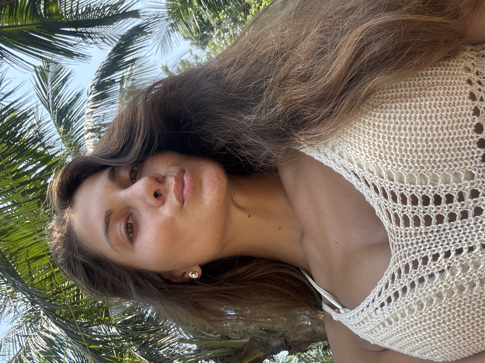 A young woman with long brown hair, fair skin, and a white knit top is pictured from a low angle against a backdrop of palm trees and a blue sky. The woman is wearing an earring. The location cannot be determined as Đà Nẵng specifically.