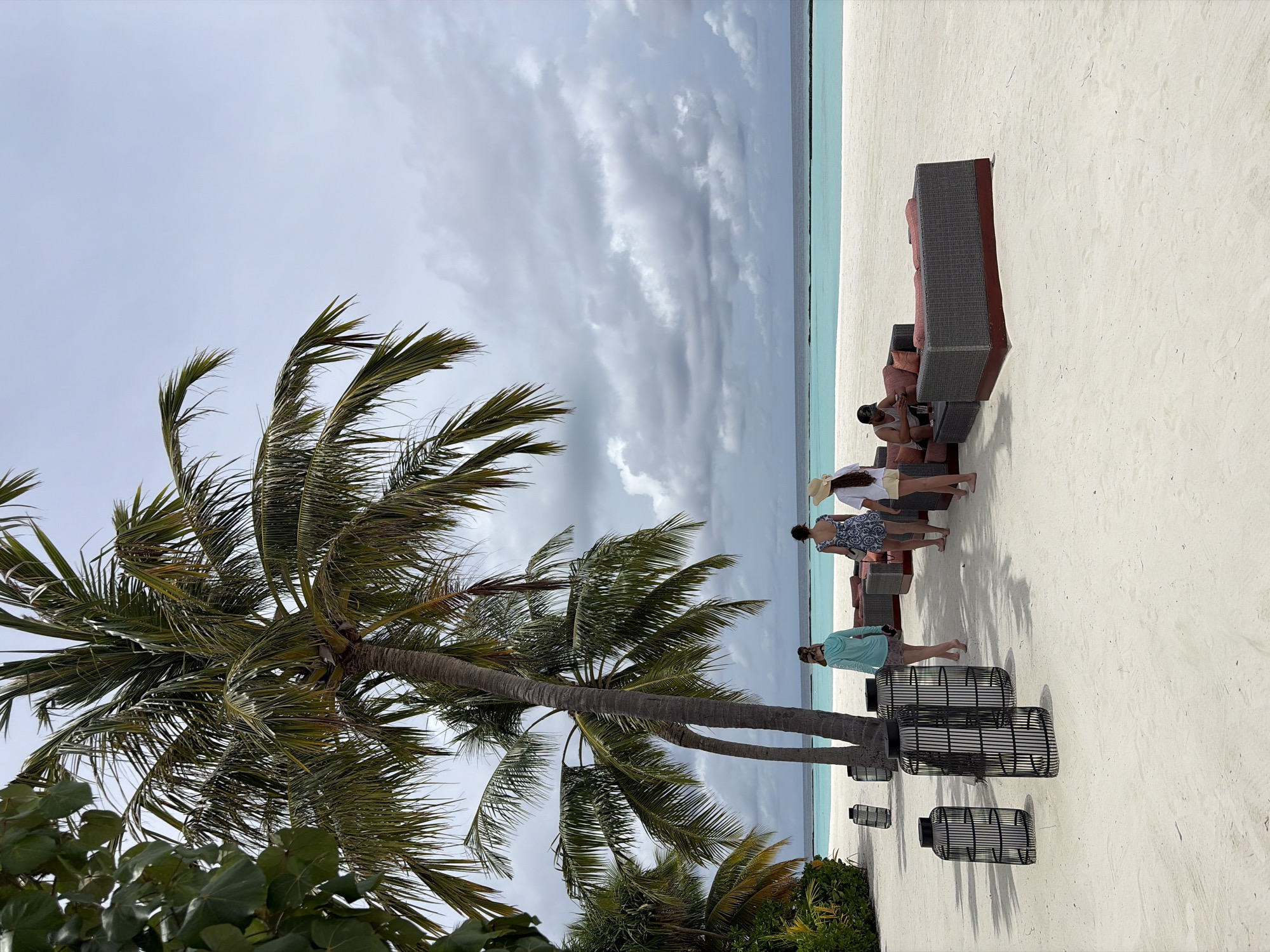 The image depicts a beach scene in Halaveli, Maldives. In the foreground, several black, cylindrical lanterns with intricate metalwork are arranged on the white sand. Beyond the lanterns, three women are walking towards the ocean. The woman on the left wears a teal hooded jacket and patterned shorts. The middle woman is in a blue and white patterned dress and a straw hat. The woman on the right wears a white top and yellow shorts with a straw hat. Further into the scene, two dark grey outdoor sofas with reddish-brown cushions are positioned on the sand. A man is seated on one of the sofas, looking at his phone. In the background, tall palm trees with lush green fronds frame the view. The turquoise ocean stretches to the horizon, meeting a cloudy, grey sky. The beach is composed of fine white sand.