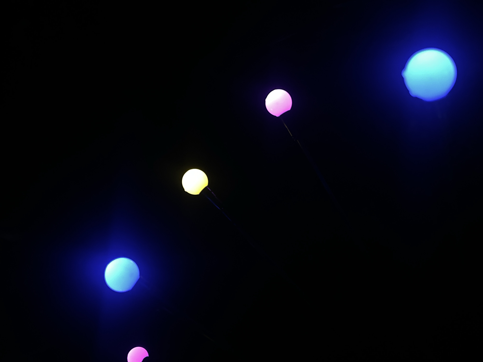 Five illuminated spheres of varying colors are sparsely distributed across a black background. The spheres appear to be attached to thin supports. The colors include blue, yellow, and pink. The arrangement is asymmetrical. The context of Novopokrovskaya, Russia, is not explicitly visually represented.
