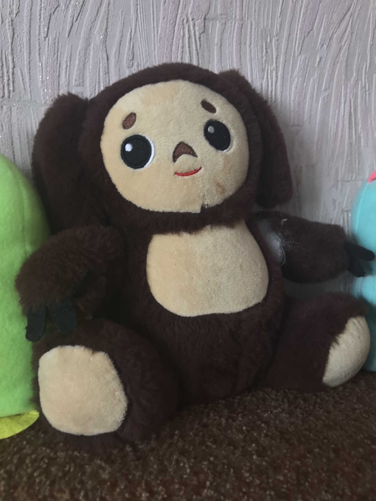 A dark brown plush toy, featuring a beige face, chest, and paw pads, is centrally positioned, seated upright on a textured dark brown carpet. The toy presents large black oval eyes with white pupils, a small brown oval nose, and a red stitched smile. A faint discolored mark is visible on its face, below the right eye. To the immediate left, a portion of a bright green plush object is visible. To the right, a segment of a light blue plush object with a pink element is discernible. The background consists of a light-colored, vertically textured wall, exhibiting small reflective glints. The scene is located in the city of Sarapul, Russia.