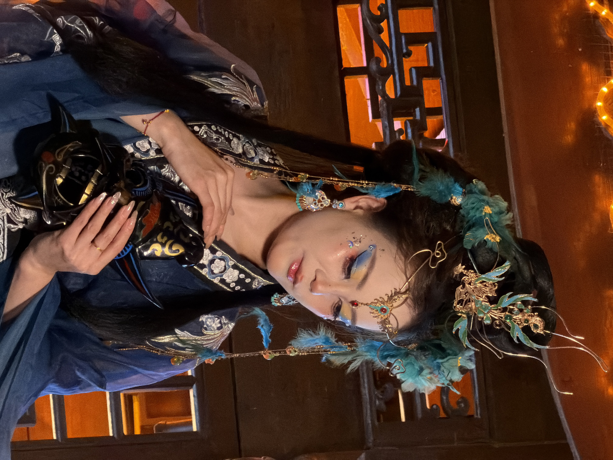 An adult East Asian female, dressed in a blue, traditional Chinese-style costume with intricate silver embroidery and embellishments, is pictured from the chest up, looking downwards. She wears an ornate headdress with teal feathers and gold accents, and elaborate makeup featuring blue and yellow eyeshadow, glitter on her cheeks, and a red gem on her forehead. Dangling earrings with blue feathers and beads adorn her ears, and a thin gold ring is visible on her left ring finger. She is holding a black, stylized mask with gold detailing in both hands. The background is a dimly lit, wooden interior with an orange-tinted lattice pattern visible through what appears to be a doorway or screen. The lighting suggests an indoor setting, possibly a studio or traditional Chinese architecture.