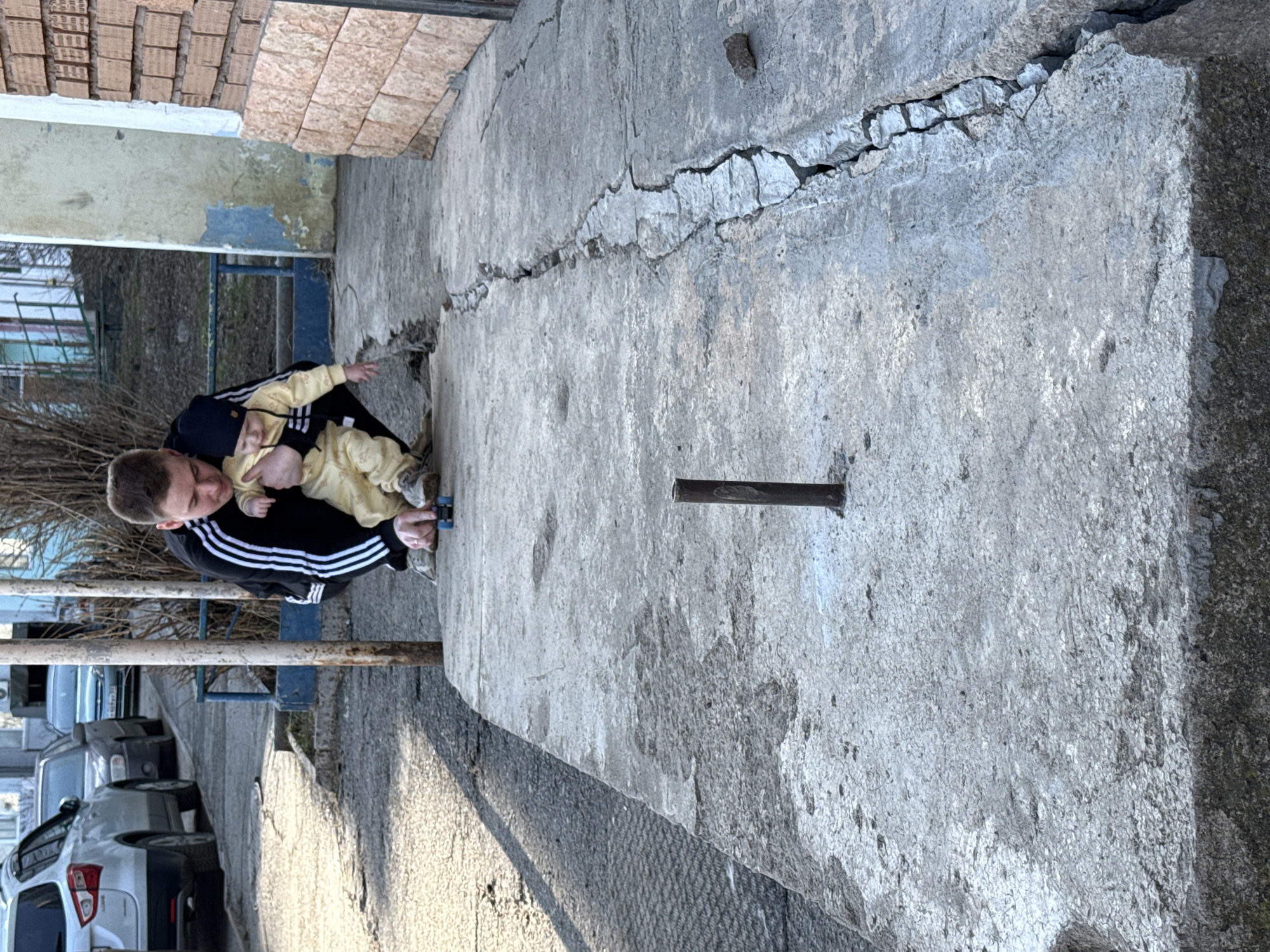 A man is crouching on a wide, cracked concrete surface next to a building. He is holding a baby in a yellow outfit. The man is wearing a black jacket with white stripes on the sleeves. The location is Chaikovsky, Russia. Cars are visible parked on the street to the left. There is a metal railing and leafless trees in the background. The concrete surface has a large crack running through it. There is a metal rod sticking out of the concrete.