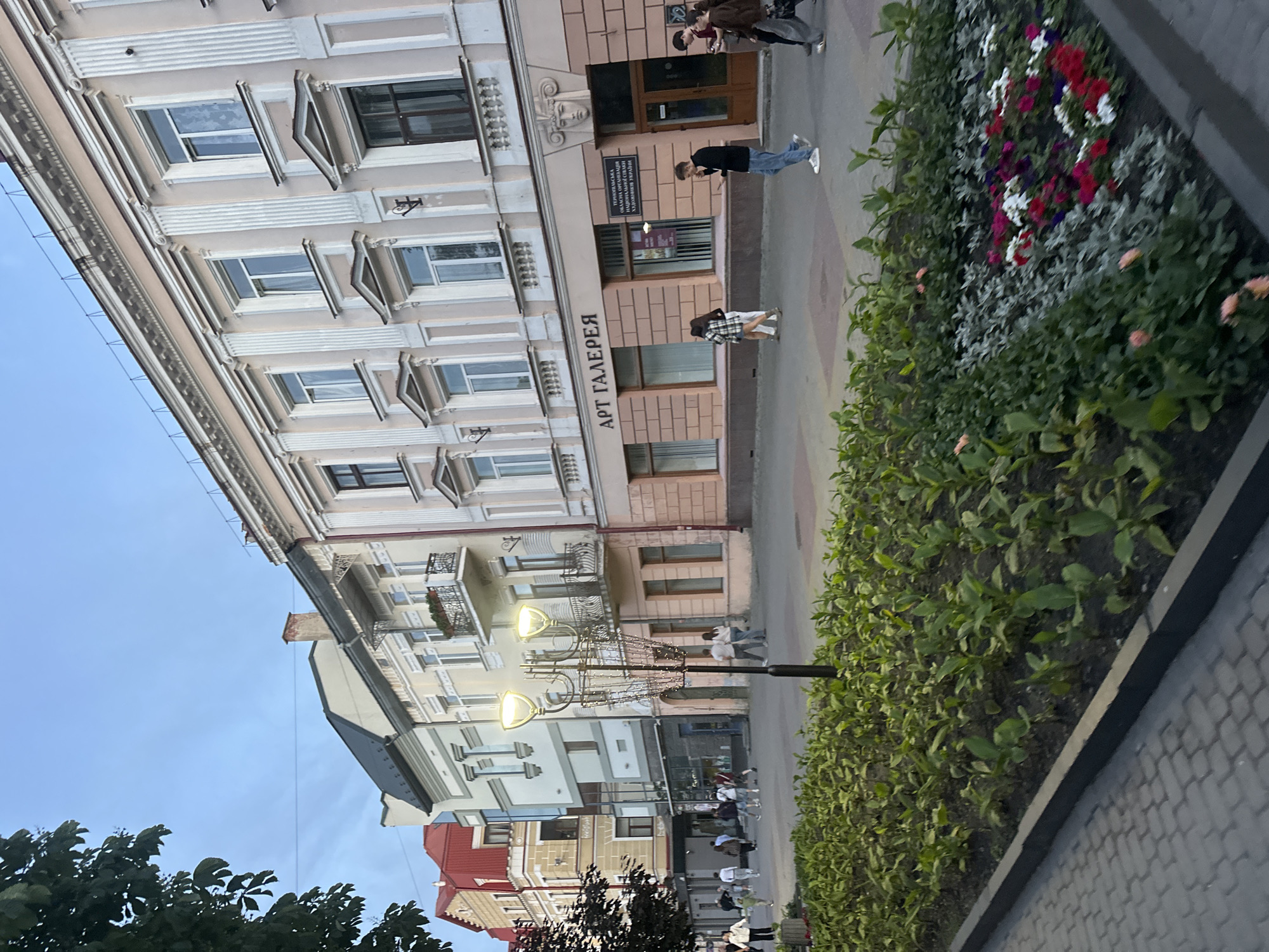The image depicts a pedestrian street in Ternopil, Ukraine, specifically in front of the "АРТ ГАЛЕРЕЯ" (Art Gallery), which is also identified by a sign for the "Тернопільська обласна організація національної спілки художників України" (Ternopil Regional Organization of the National Union of Artists of Ukraine).
The scene features a row of multi-story buildings lining the left side of the street, viewed from a slight angle. The prominent building on the right, housing the art gallery, is light peach-colored with classical architectural elements, including decorative columns, cornices, and window frames. A sculpted head adorns the facade above the gallery's main entrance. To its left, another building features ornate iron balconies and a lighter cream facade. Further down the street, buildings include modern sections with glass and older structures, one distinguished by a red-tiled roof at the far left. Overhead wires are visible against the pale blue sky.
The street itself is paved with grey interlocking pavers. A central elongated garden bed runs along the pedestrian path, planted with various green foliage. Towards the right side of the garden bed, sections contain mixed plantings, including silver-leafed plants and clusters of red, white, and purple flowers. A tall, decorative street lamp with multiple arms and what appear to be string lights is centrally positioned within the garden bed.
Multiple pedestrians are observed walking along the street in various directions. Most individuals are casually dressed. On the right side, a man in a black long-sleeved shirt and blue jeans walks while looking at a phone. Behind him, two other individuals also appear to be looking at phones. Towards the center-left, several groups of people are walking, some interacting, others individually. A few individuals are observed standing near shopfronts. Tree foliage is visible in the upper left corner of the frame.