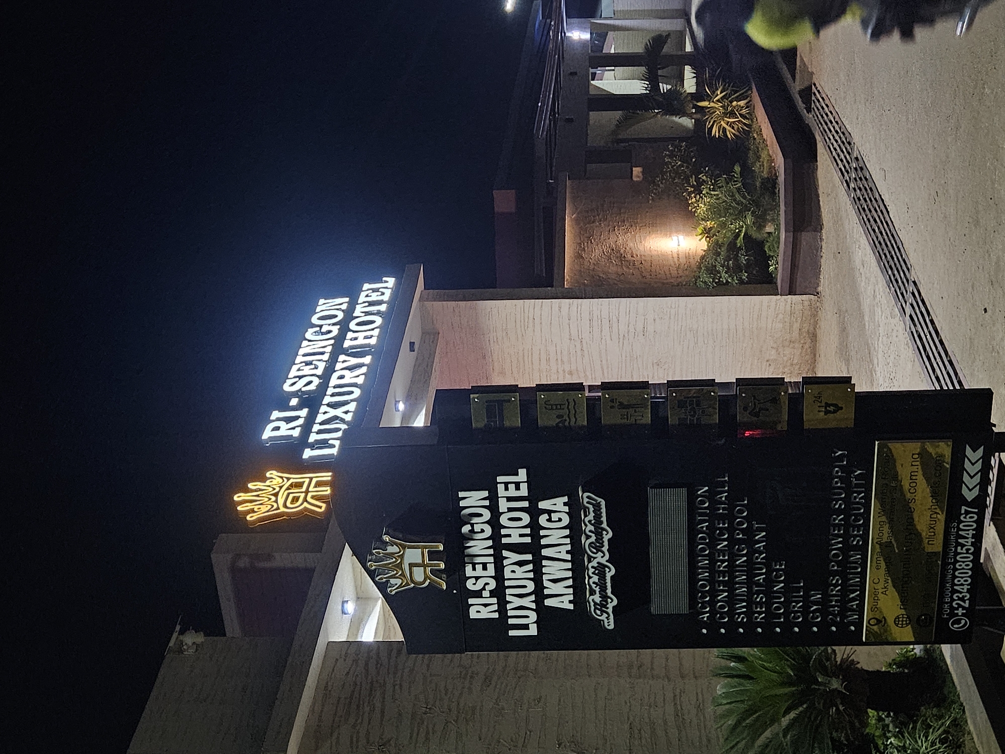 The image is a nighttime view of the "RI-SEINGON LUXURY HOTEL" in Akwanga, Nigeria. The hotel name is prominently displayed on a sign in bold, illuminated letters. A smaller sign lists amenities such as accommodation, a conference hall, swimming pool, restaurant, lounge, grill, gym, 24-hour power supply, and maximum security. The hotel is located on Super C ema, Along Warriba Road, Akwanga, Nassarawa State. The website addresses riseingonluxuryhotes.com.ng and nluxuryhotels.com are also listed. A phone number for bookings is provided: +2348080544067. The surrounding environment appears to be urban, with some greenery visible near the building.
