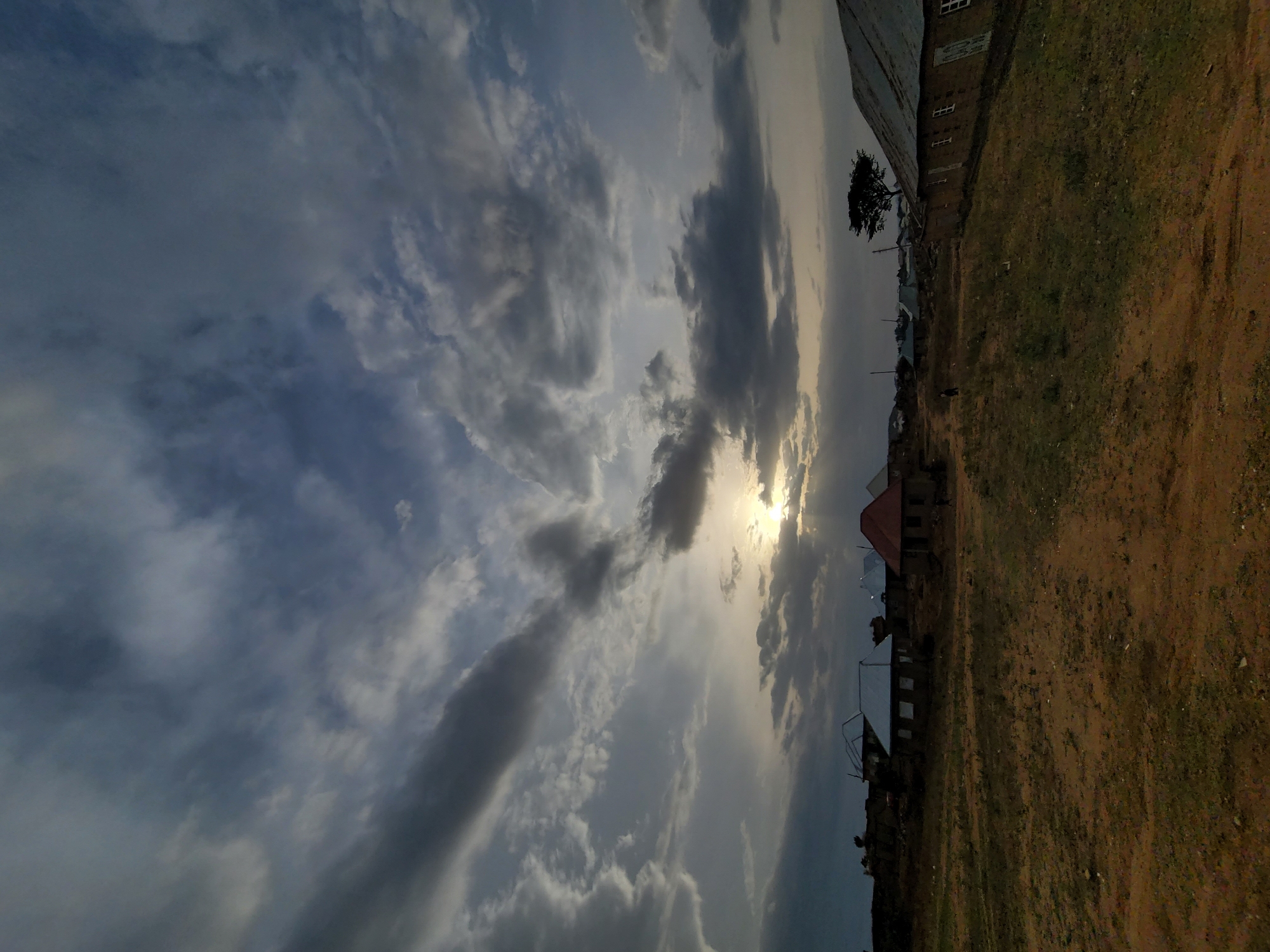 This outdoor landscape photograph captures a serene scene under a vast sky filled with dramatic clouds. The sun, partially obscured by these clouds, casts a golden and diffused light across the horizon, strongly suggesting either late afternoon or early evening. The ground, which occupies the lower half of the image, is characterized by dry, reddish-brown earth with sparse patches of green vegetation.
In the midground and background, several simple structures are visible, including buildings with what appear to be corrugated metal roofs. One building stands out with a dark, possibly red, roof. A solitary tree with dark foliage is silhouetted against the bright sky near these structures. No people or specific activities are depicted in the image, contributing to a calm and expansive atmosphere. The earthy terrain and functional architecture align with a rural or semi-urban environment, consistent with the Barakin Akawo, Nigeria location. There is no visible text.