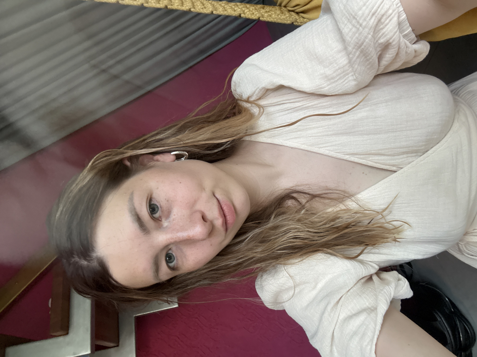 A young woman with light brown hair and blue eyes is posing for a selfie. She is wearing a light-colored blouse. The background includes a burgundy surface, a gray striped surface, and a gold rope. The location in the city of Чёрное, Russia is not discernible from the provided image.