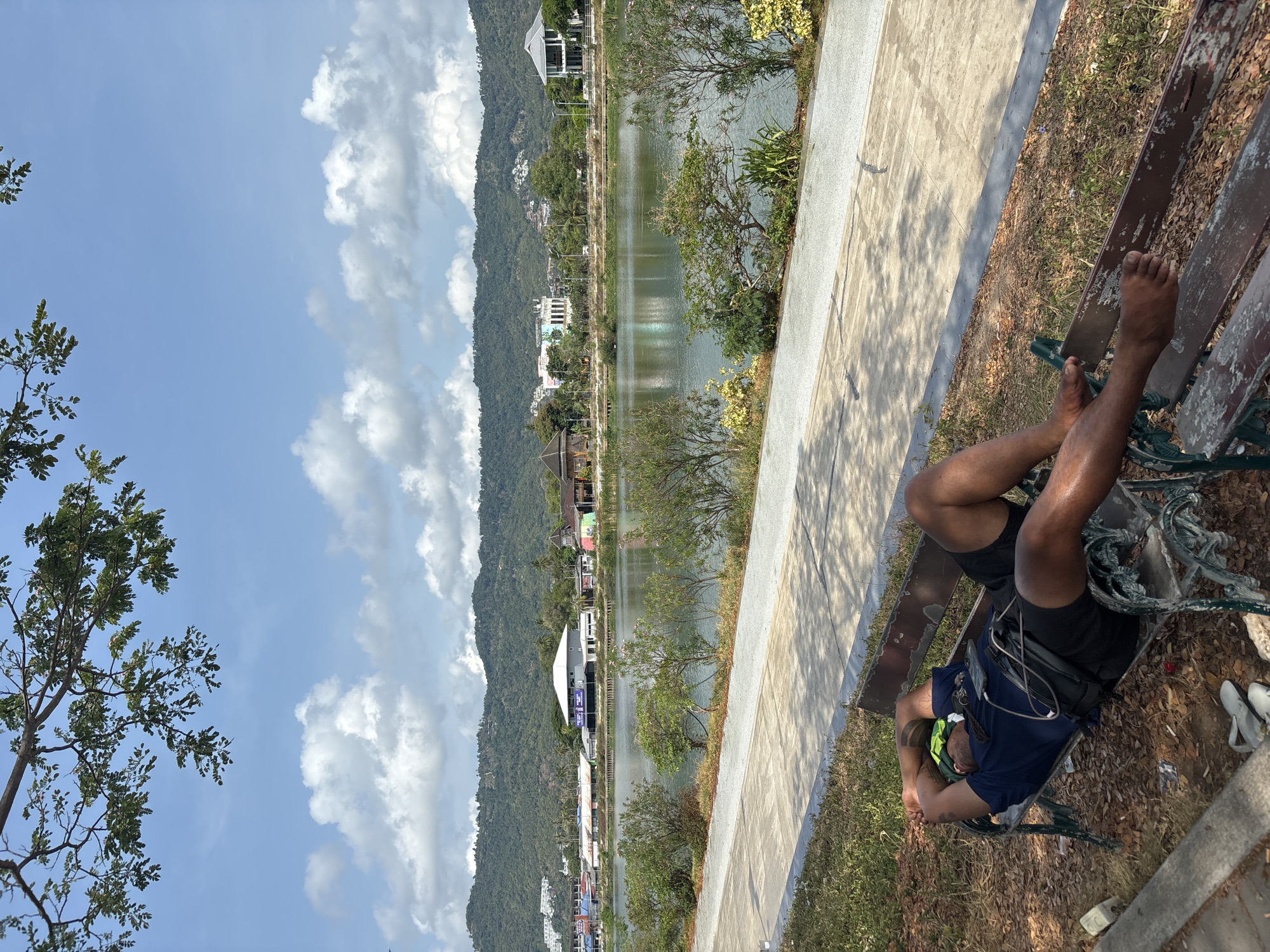 The image shows a man sleeping on a park bench in Baan Chaweng, Thailand. He is lying on his back with his feet propped up on the back of the bench. He is wearing a dark blue shirt, black shorts, and a fanny pack. His head rests on his folded arms. A neon green object is visible near his head. The bench is metal and weathered. Next to the bench is a concrete walkway, with a lake and mountains in the background. The sky is blue with puffy white clouds. Some buildings can be seen along the edge of the lake and at the base of the mountains.
