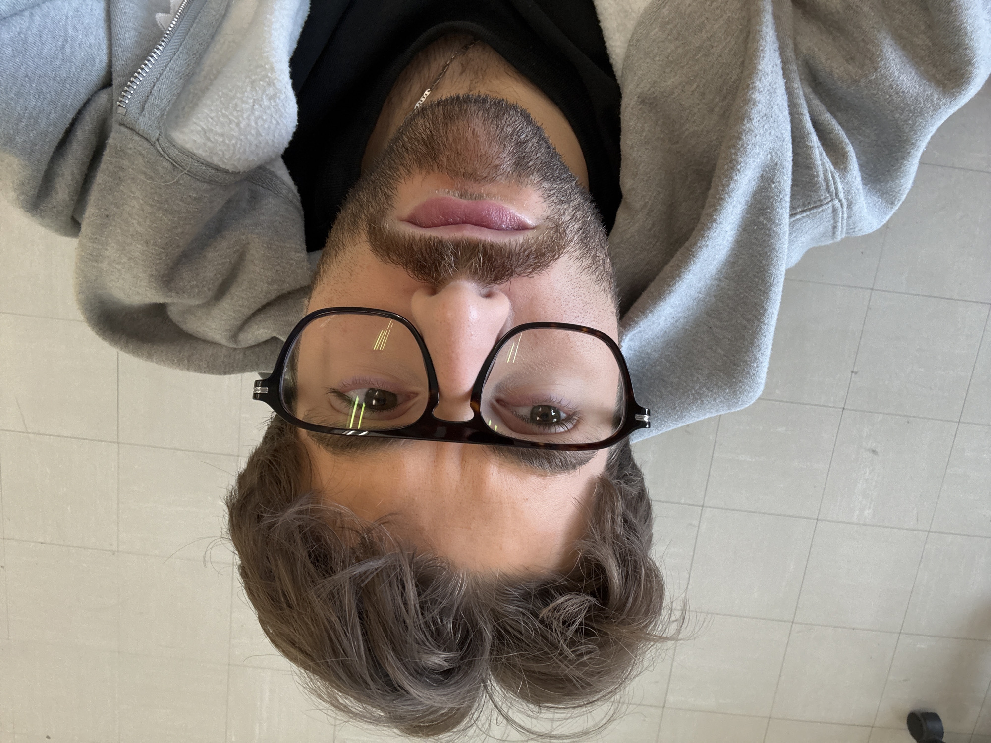 The image displays an upside-down close-up portrait of a young man, appearing to look up towards the camera. His head, featuring light brown, slightly wavy hair, is positioned at the bottom of the frame. He has a neatly trimmed beard and mustache and wears dark-framed glasses over his eyes, which are visible at the lower part of his face. His lips are slightly parted, and his expression is neutral.
He is dressed in a grey, fleece-lined hooded sweatshirt, which includes a prominent zipper detail, worn over a dark garment visible beneath. Reflections are noticeable on the lenses of his glasses. The background consists of a light-colored tiled floor, suggesting an indoor setting. There is no discernible text present in the image. The provided context indicates the location is Montreal, Canada.