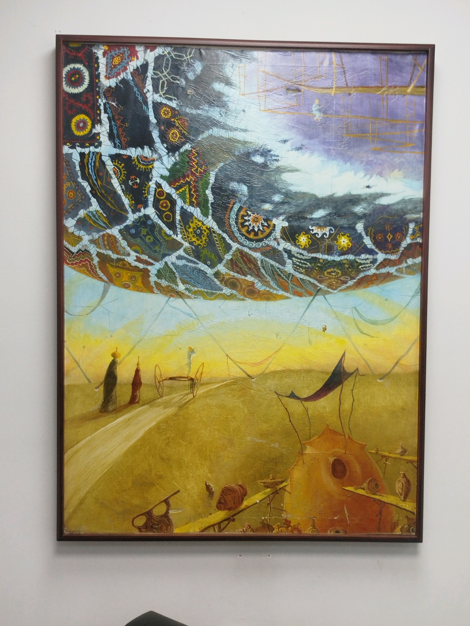 This is a photograph of a painting framed in dark wood, hanging on a white wall. The painting itself depicts a surreal, dreamlike landscape.
In the upper portion of the painting, a vast, fragmented canopy hangs in a purple and light blue sky. This canopy appears to be made of colorful, intricately patterned fabric, resembling traditional Uzbek textiles, pieced together with white stitching. Some parts of the canopy are dark and cloudy, while others are illuminated. Golden, lattice-like structures are visible within the sky, possibly suggesting scaffolding or ethereal architecture.
The lower half of the painting is a golden-yellow landscape, suggesting a desert or a dry plain. A pale yellow path winds through this landscape. On this path, three figures are present. The two figures in the foreground are dressed in long, dark and red garments, with one wearing a crown-like headdress. They appear to be walking or standing. Further along the path, a small, two-wheeled cart is depicted. A third, smaller figure stands near the cart.
In the middle ground and foreground, various abstract and organic structures are scattered across the landscape. A prominent, orange, dome-shaped object with a circular opening is situated on the right. This structure is supported by thin, dark poles and has yellow platforms extending from it, on which small, ceramic-like objects are placed. Other smaller, sculptural elements and what appear to be tools or machines are also visible in this area. The overall impression is one of a fantastical and somewhat unsettling scene.
The style of the painting is highly imaginative and detailed, with a focus on intricate patterns and symbolic representation. The artwork is characteristic of the surrealist movement.
This painting is located in the Museum of Applied Arts of Uzbekistan, in Tashkent, Uzbekistan. The museum itself is housed in a historic building that was once the residence of diplomat Alexander Polovtsev.