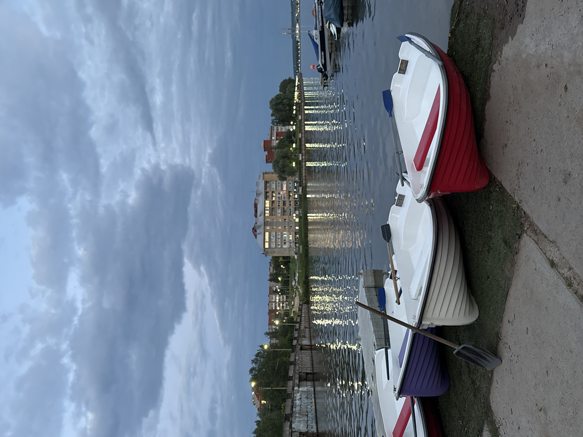 The image depicts a waterfront scene in Votkinsk, Russia, likely during twilight. In the foreground, several small rowing boats are pulled ashore on a grassy bank adjacent to a concrete walkway. One boat is bright red, another is white, and a third is purple. A shovel rests against the purple boat. In the middle ground, the calm water of a river or lake reflects the lights from buildings and streetlamps along the opposite shore. A concrete pier or embankment runs along the left side of the frame. Beyond the water, a line of multi-story buildings with illuminated windows stretches across the landscape, interspersed with trees. The sky is overcast with heavy, dark clouds, suggesting an approaching storm or the end of the day. The overall atmosphere is serene but somewhat somber due to the muted lighting and cloudy sky.