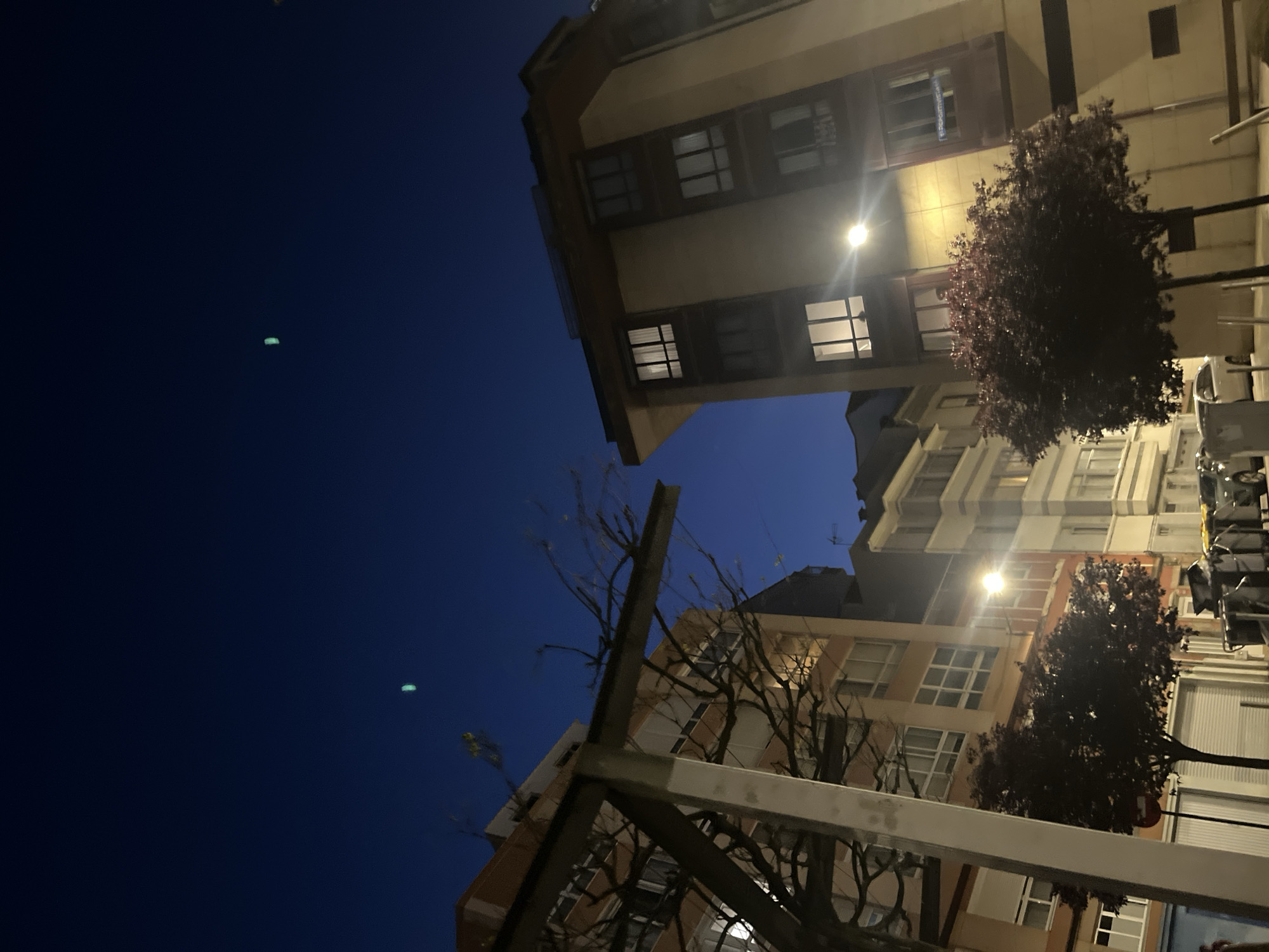 The image, taken at night, depicts a view of buildings in A Coruña, Spain. The perspective is looking upwards from street level. The buildings are a mix of beige and brown, with multiple windows visible, some illuminated. Street lights cast a yellow glow, affecting the buildings and the surrounding foliage. Trees with bare branches and trees with dense, dark-colored foliage are present. The sky is dark blue. A metal structure is visible in the foreground. There are no people visible in the image.