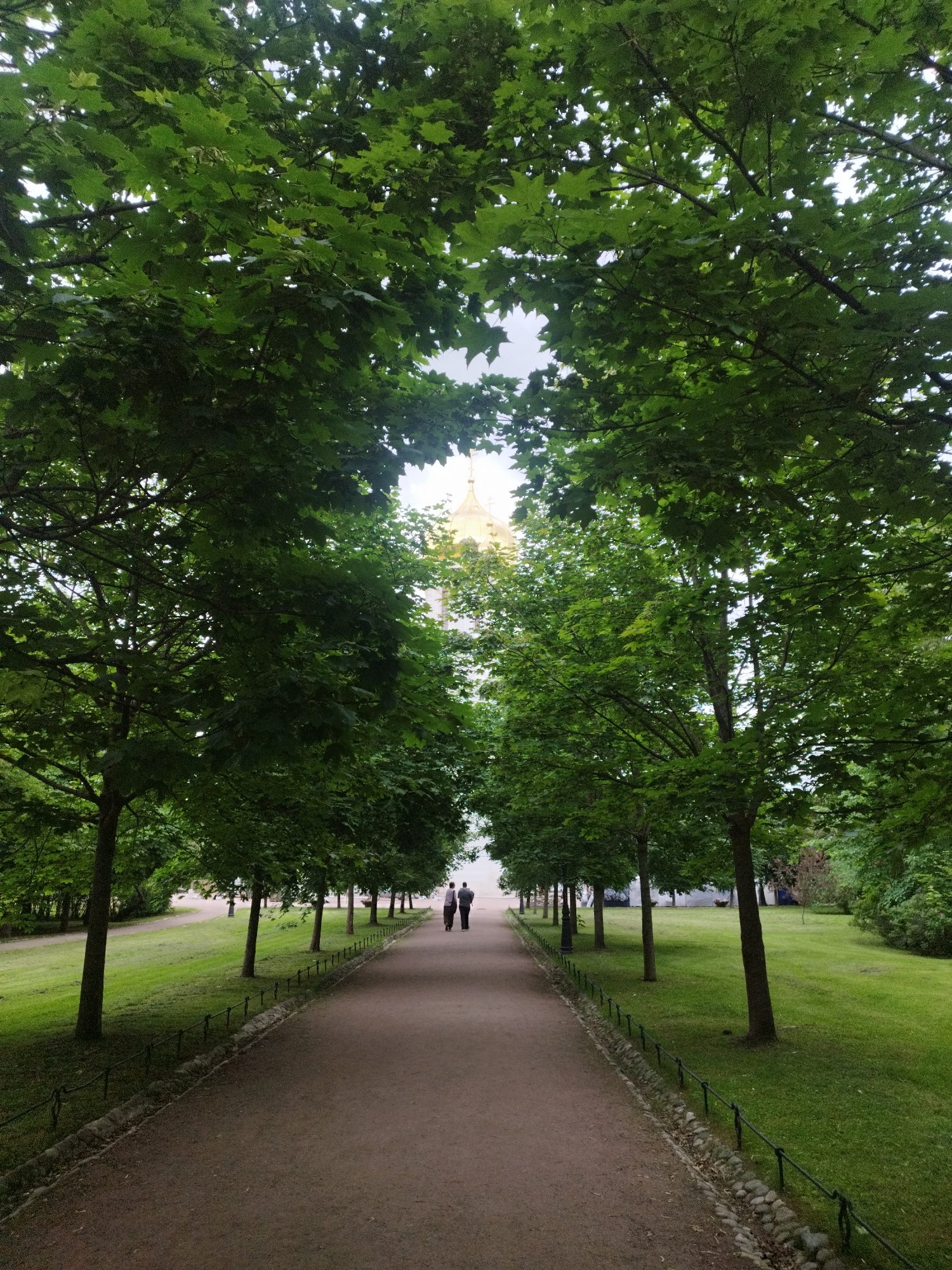 A person, or people, are walking on a dirt path lined with trees on both sides. The trees are green and leafy. In the distance, there is a golden dome visible. The ground is covered in green grass. The path is in the middle of the image and is a light brown color. There are two people walking on the path away from the camera. The trees create a tunnel-like effect over the path. The sky is not visible due to the trees. The lighting suggests it is daytime.