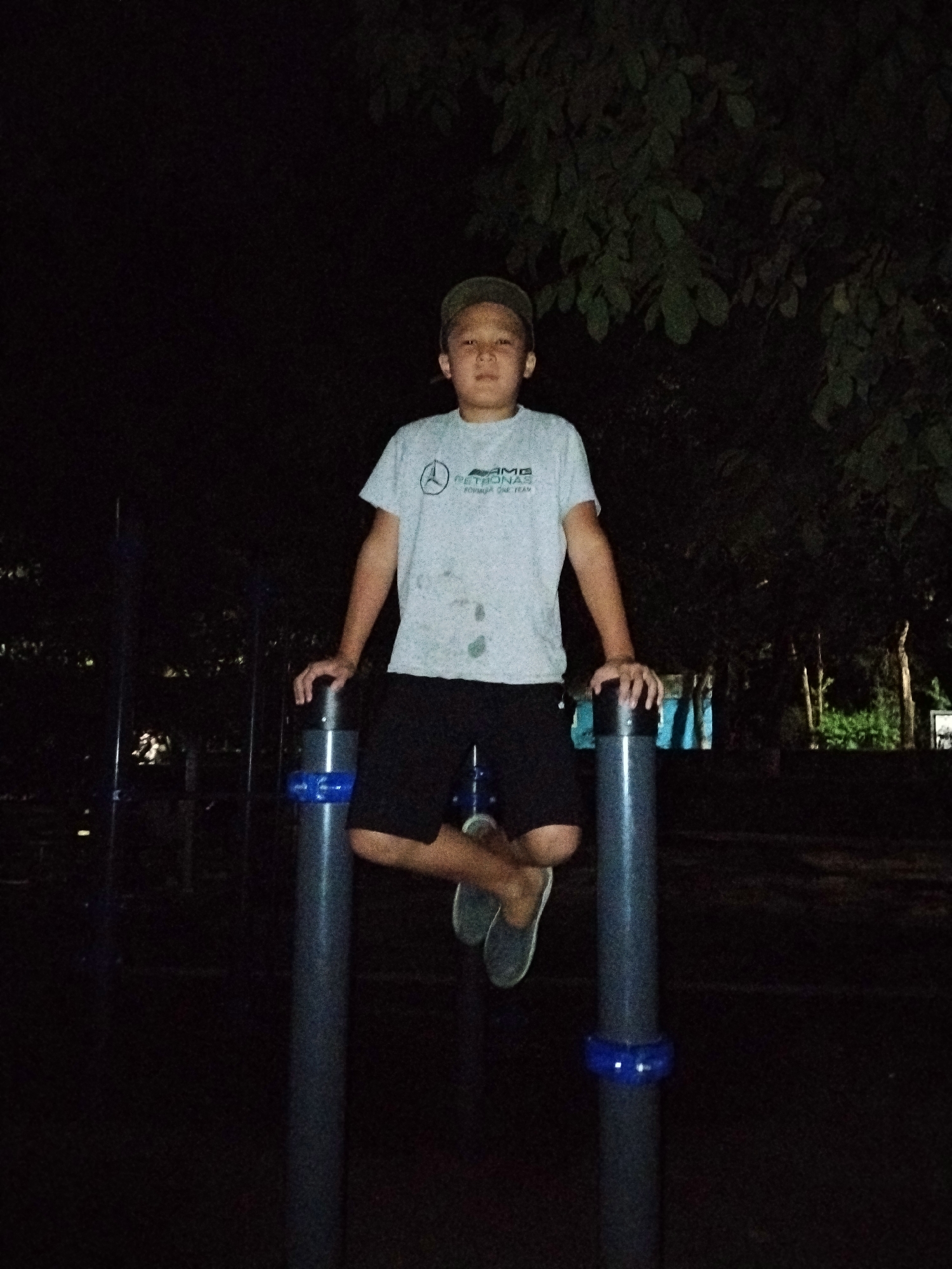 In Bishkek City, Kyrgyzstan, at night, a young boy wearing a light gray t-shirt with a logo, dark shorts, a green cap, and gray slip-on shoes is positioned between two tall, dark gray poles with blue bands. The boy is sitting on the poles, with his legs bent and crossed. His right hand is resting on the left pole, and his left hand is on the right pole. The background is dark, with some foliage visible at the top and scattered lights in the distance, suggesting an outdoor location, possibly a park or playground.