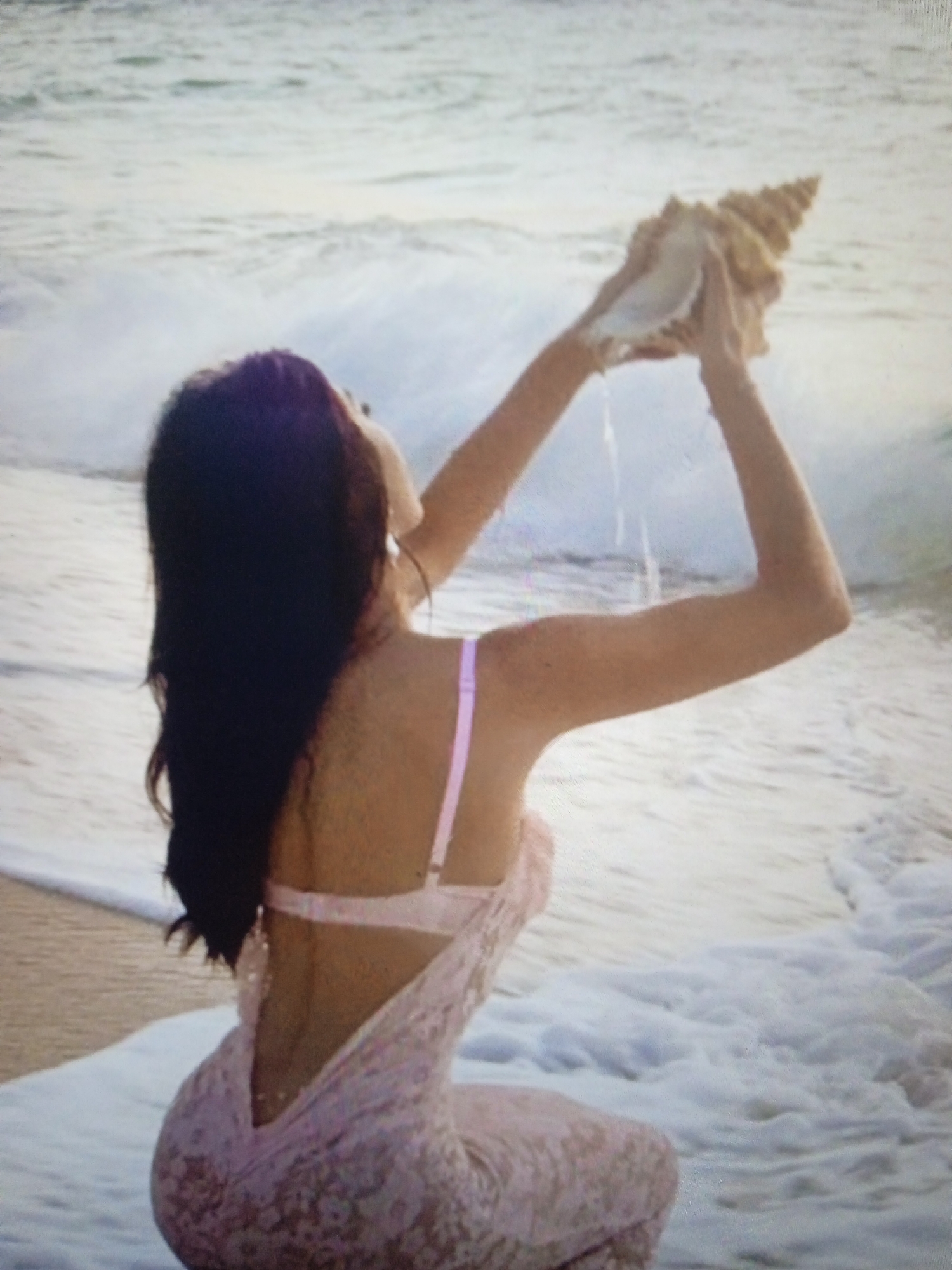 A woman is sitting on a beach holding a large seashell up to her ear, with waves crashing around her. She has long dark hair and is wearing a light pink dress with a low back. The water is a pale blue and white, and the sand is a light brown. There is no indication of this image being taken in Chernivtsi, Ukraine.