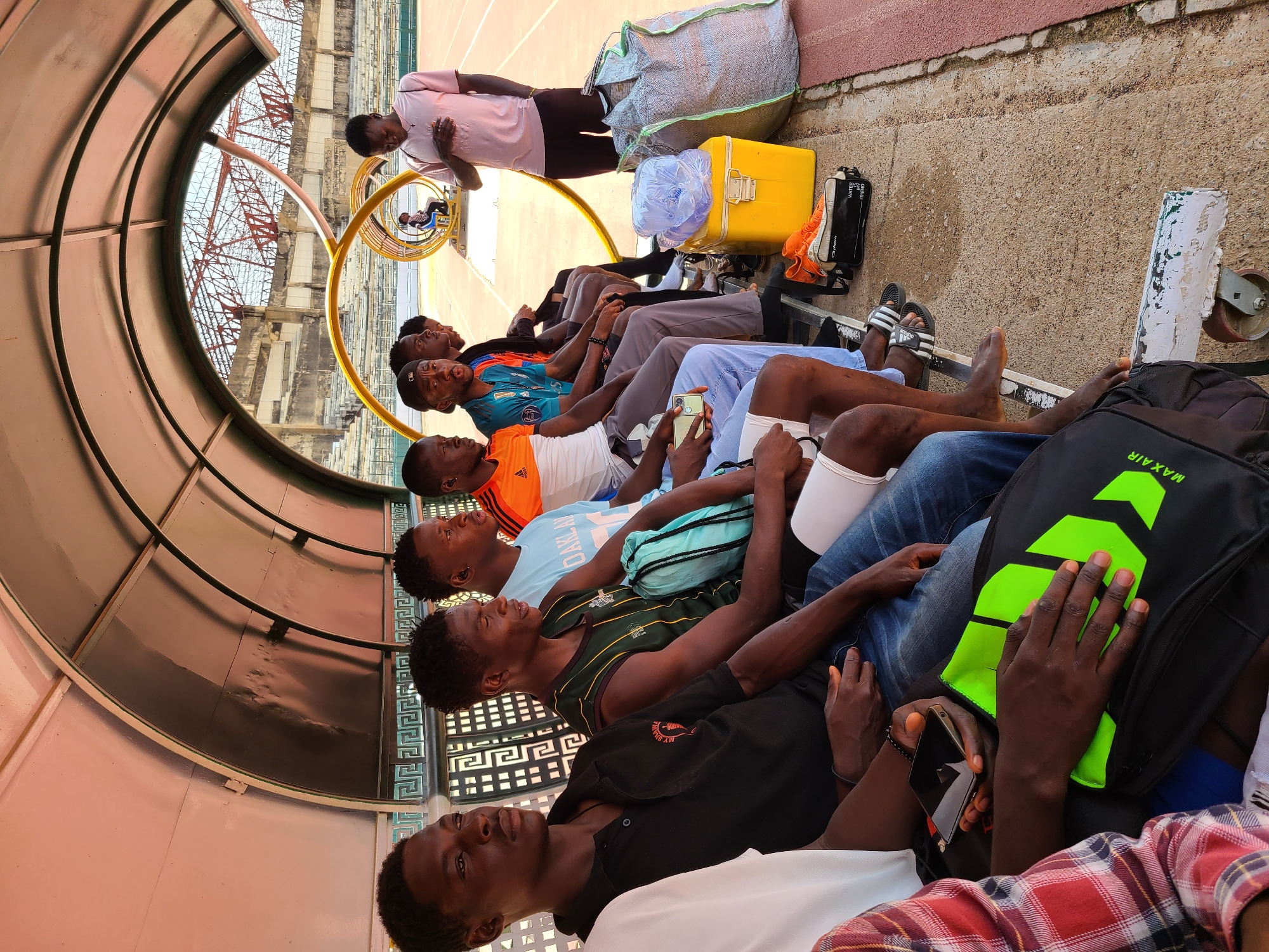 The image shows a group of approximately ten young men sitting under a sheltered area at what appears to be a track and field stadium. The men are diverse in their attire, some wearing athletic clothing and others in casual wear like jeans and button-down shirts. Some of them are looking forward while others appear to be distracted by cellphones. Various personal belongings like a yellow cooler, bags (including one branded "MAX AIR"), shoes, and water bottles are scattered beside them on the concrete surface. A single man in a pink shirt stands behind the seated men, seemingly in conversation with them. In the background, stadium seating and support structures are visible. The location is specified as the New Stadium, Nigeria.