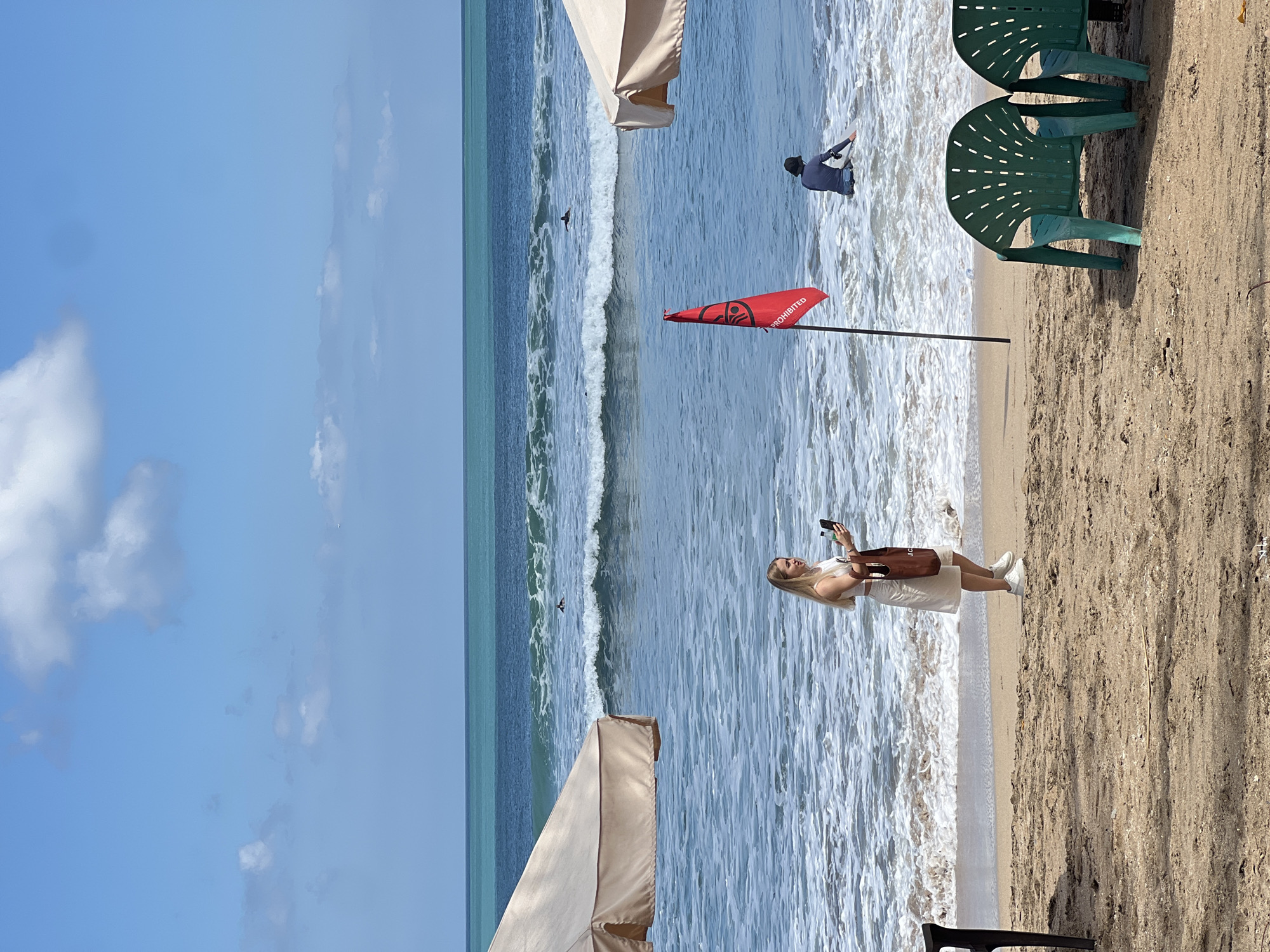 The image depicts a sunny day at Kuta Beach, Bali, Indonesia. The foreground is dominated by sandy beach with two green plastic beach chairs. In the middle ground, a woman with blonde hair wearing a white dress and white sneakers is standing by the water's edge, holding a smartphone and a brown tote bag. She appears to be taking a photo or video. A red flag with a black symbol and the word "PROHIBITED" printed vertically is planted in the sand and water nearby. In the background, the ocean displays turquoise water with white-capped waves. Several surfers are visible in the distance, some riding waves and one sitting on a surfboard. Two large, beige beach umbrellas are positioned on the sand, partially framing the scene. The sky is blue with scattered white clouds.
