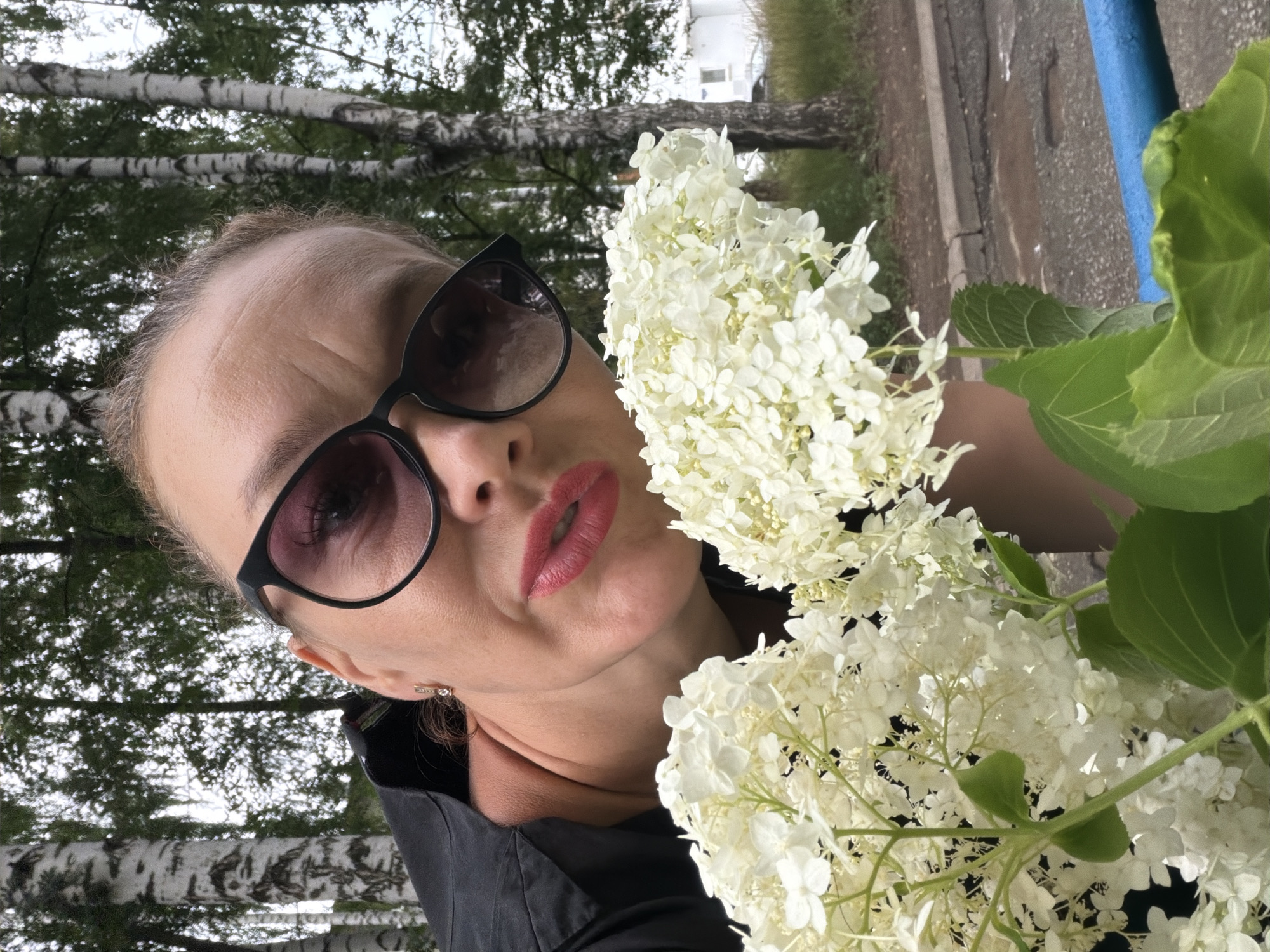 A woman wearing black clothing and sunglasses poses for a selfie outdoors, holding a bouquet of white hydrangeas. She is positioned in front of birch trees, with a building and a street visible in the background. The setting is consistent with an urban park or residential area in Chaikovsky, Russia, likely during the daytime given the natural light. The woman has bright red lipstick and appears to be looking directly at the camera with a slightly pursed expression. The overall scene is casual and personal.