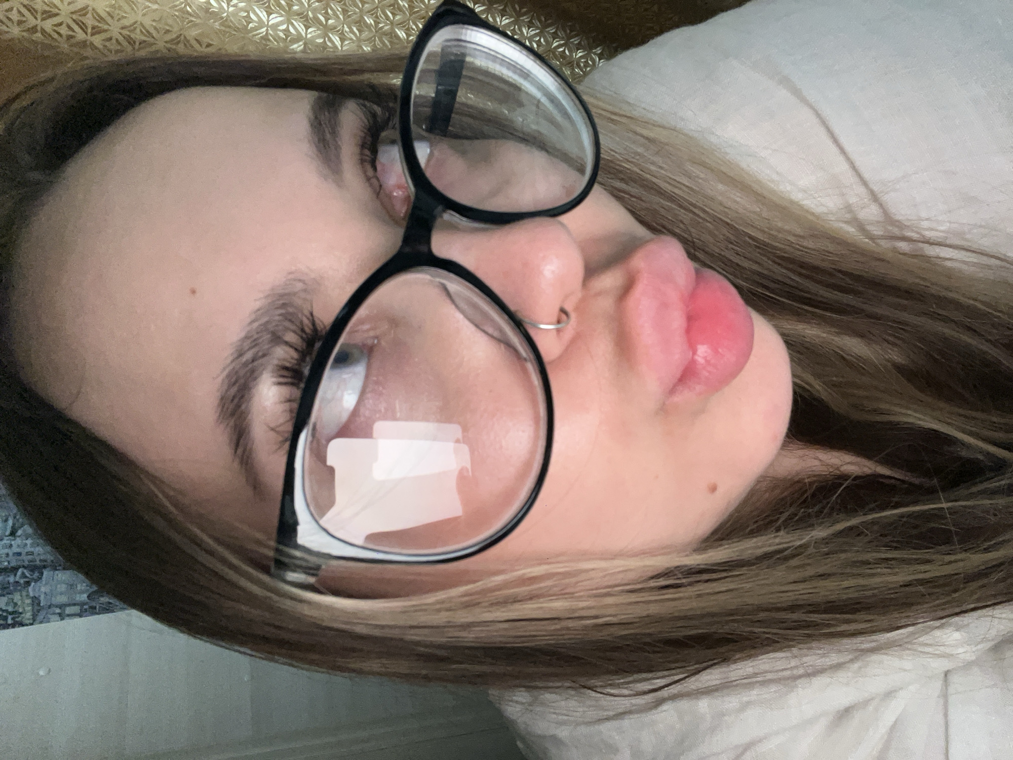 A person with light skin and brown hair wearing black-rimmed glasses is lying down. The person has a nose ring and is pursing their lips. The background includes a textured gold surface and white fabric. The location is unspecified, but the city is mentioned as Novopokrovskaya, Russia.