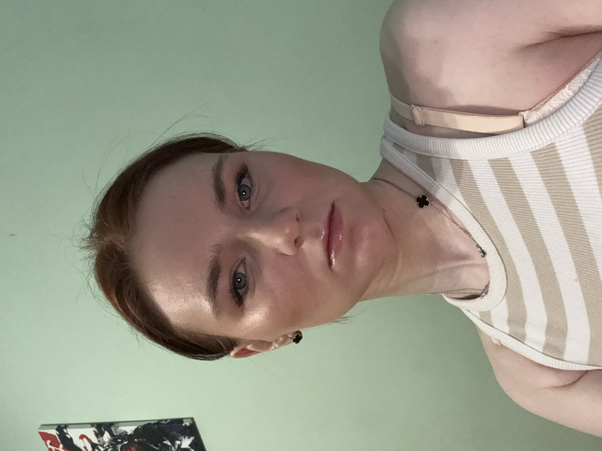 A woman with red hair and fair skin is shown in a selfie-style image. She is wearing a striped tank top and a delicate silver necklace with a small, black, four-leaf clover charm. The background is a solid light green. The image is slightly rotated counter-clockwise. A small object, partially visible in the lower left corner, appears to be a box or book with a graphic design featuring shades of red, white, and black. There is no information in the image to indicate that this image was taken in Vsevolozhsk, Russia.