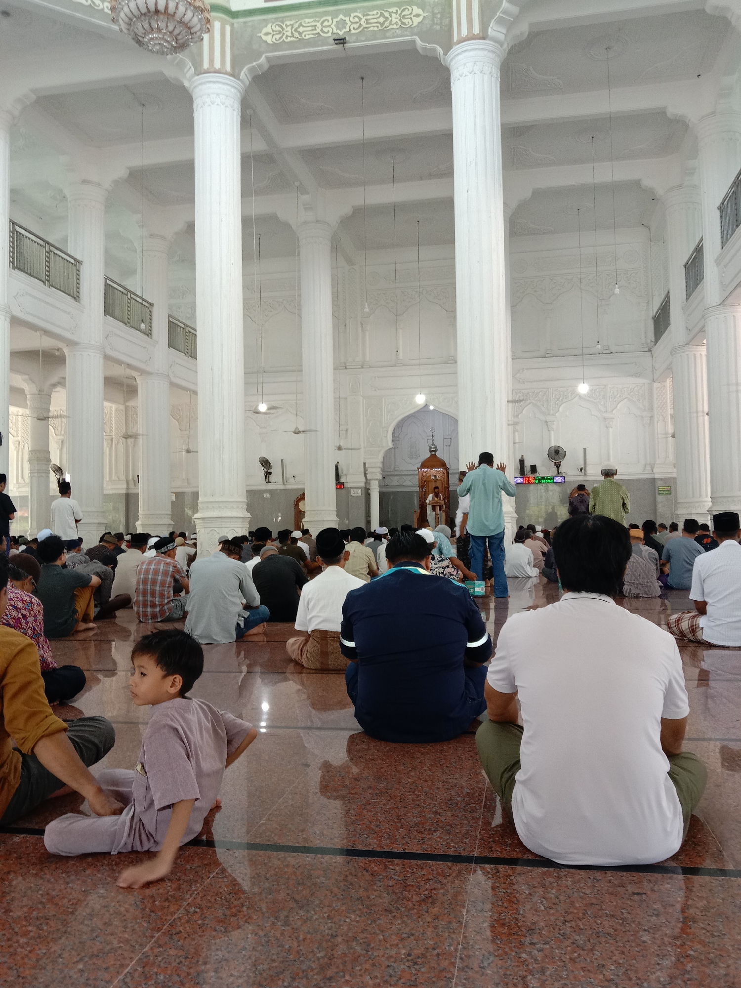 The image captures the interior of a mosque in Lhokseumawe, Indonesia, featuring a large group of people seated on the floor. The building's architecture is characterized by numerous white columns, ornate ceilings, and a chandelier. A young child sits near the front, while an individual stands near a decorated niche at the far end of the room. The setting suggests a religious gathering or prayer service. The floor is a polished, reddish-brown color. A digital clock is visible on the wall near the niche.