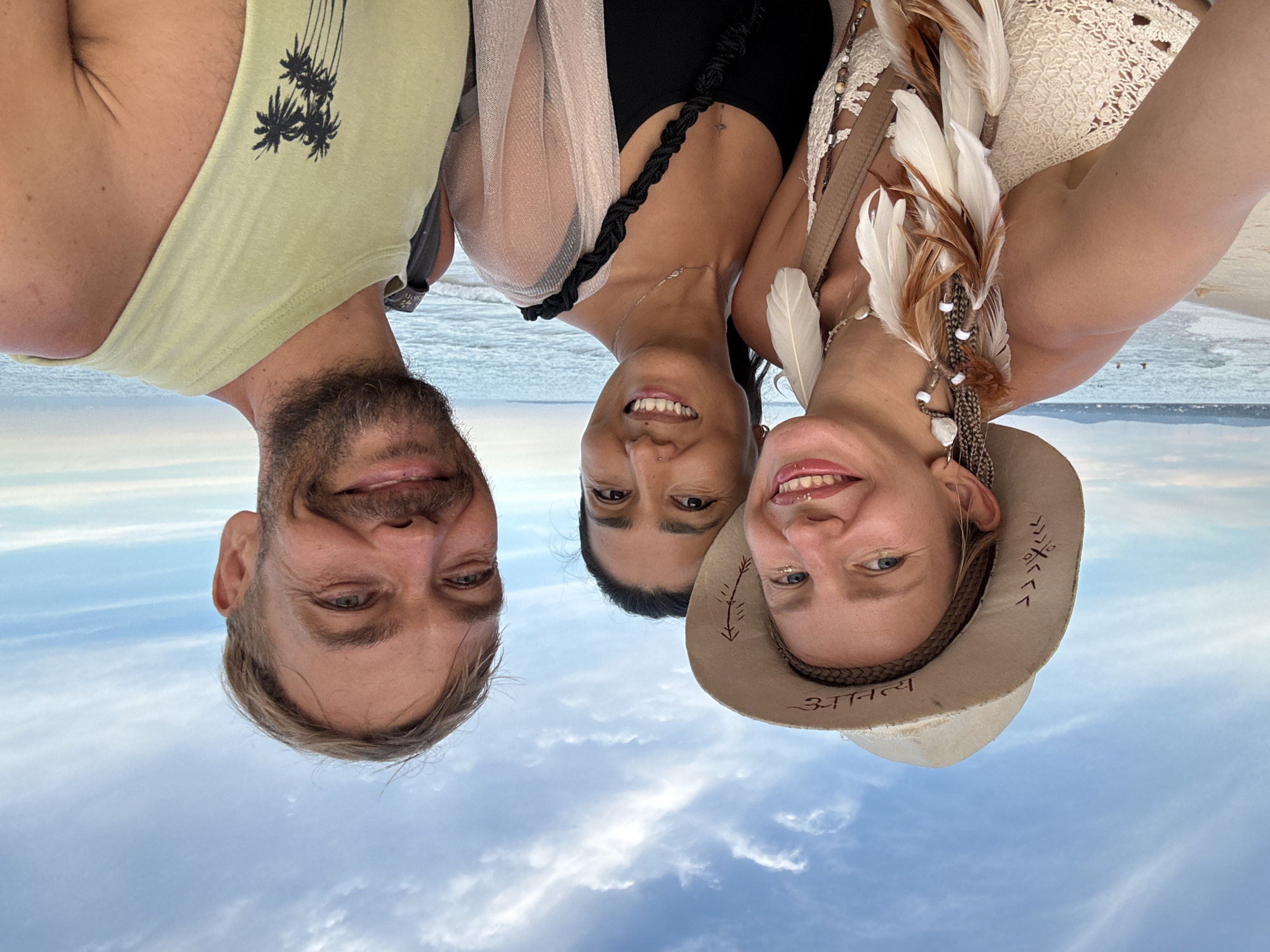 Three individuals, two women and one man, are positioned in the foreground, taking a selfie on a beach. The woman on the left wears a wide-brimmed hat adorned with feathers and intricate braided hair extensions. She has glitter on her cheeks and smiles at the camera. The woman in the center is smiling and wearing a black tank top and a sheer, sparkly cardigan. The man on the right is also smiling, with a beard and short hair, wearing a light green tank top with a palm tree design. Behind them, the ocean meets the sky, with faint clouds visible. The beach has sand in the foreground, and a body of water with a few distant figures swimming. The overall setting suggests a relaxed, tropical atmosphere.