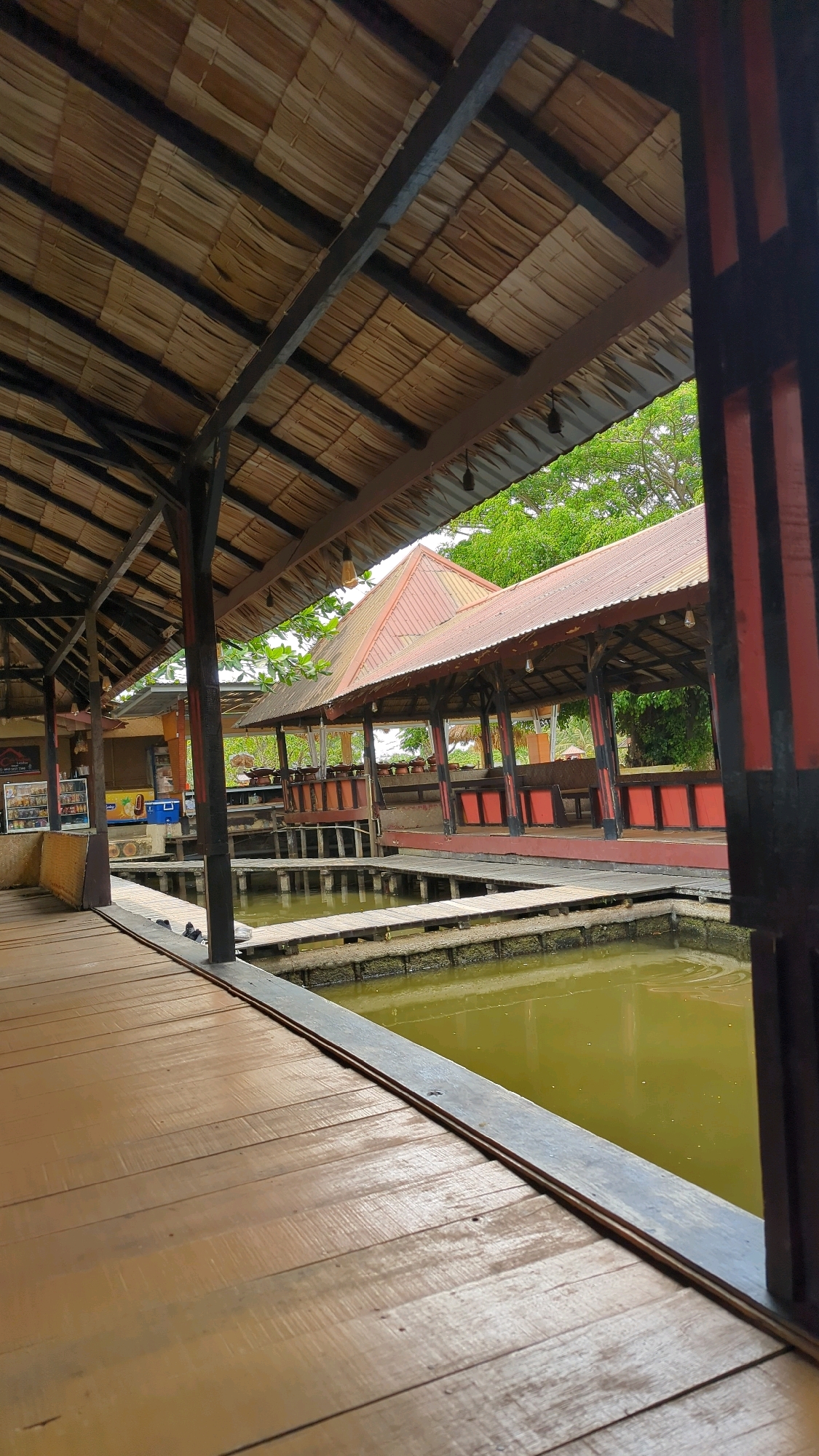 The image shows an outdoor seating area constructed over a body of water in Sungaidua, Indonesia. The structure features a thatched roof supported by dark wooden beams and columns. A wooden walkway extends from the foreground towards the seating area, which is elevated above the greenish water. The seating area has a reddish-brown roof and benches. Trees and foliage are visible in the background. The scene appears to be part of a restaurant or recreational facility. There are no people visible in the image.