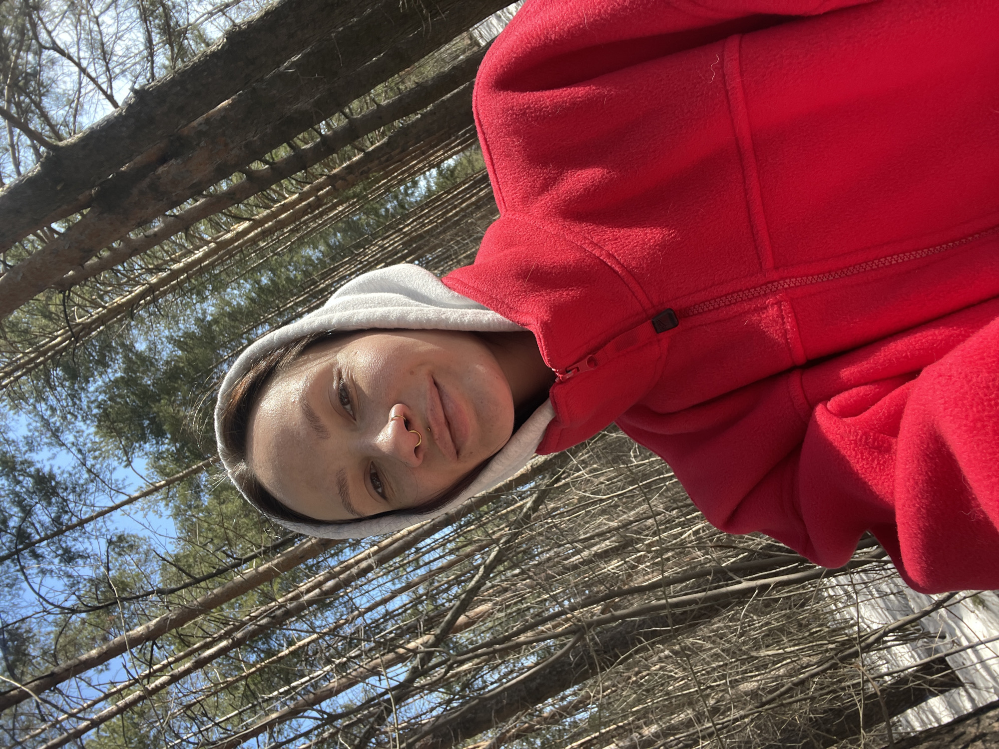 A person with a nose ring, wearing a red fleece jacket with a white hood, is posing for a selfie. The background consists of bare tree branches against a bright blue sky, suggesting a forest or wooded area. The trees appear to be deciduous, as they lack leaves. The person's face is tilted towards the camera, and the overall lighting is bright. The exact location within Izhevsk, Russia cannot be determined from the image.