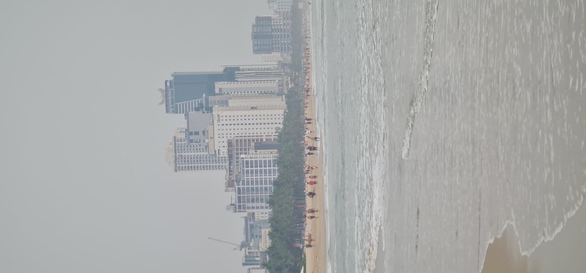 The image shows a beach in Đà Nẵng, Vietnam. Tall buildings line the coast. A sandy beach is visible with numerous people walking along the shoreline. The sea is slightly choppy. The sky is overcast, contributing to a somewhat hazy atmosphere. Palm trees line the boundary between the beach and the buildings.